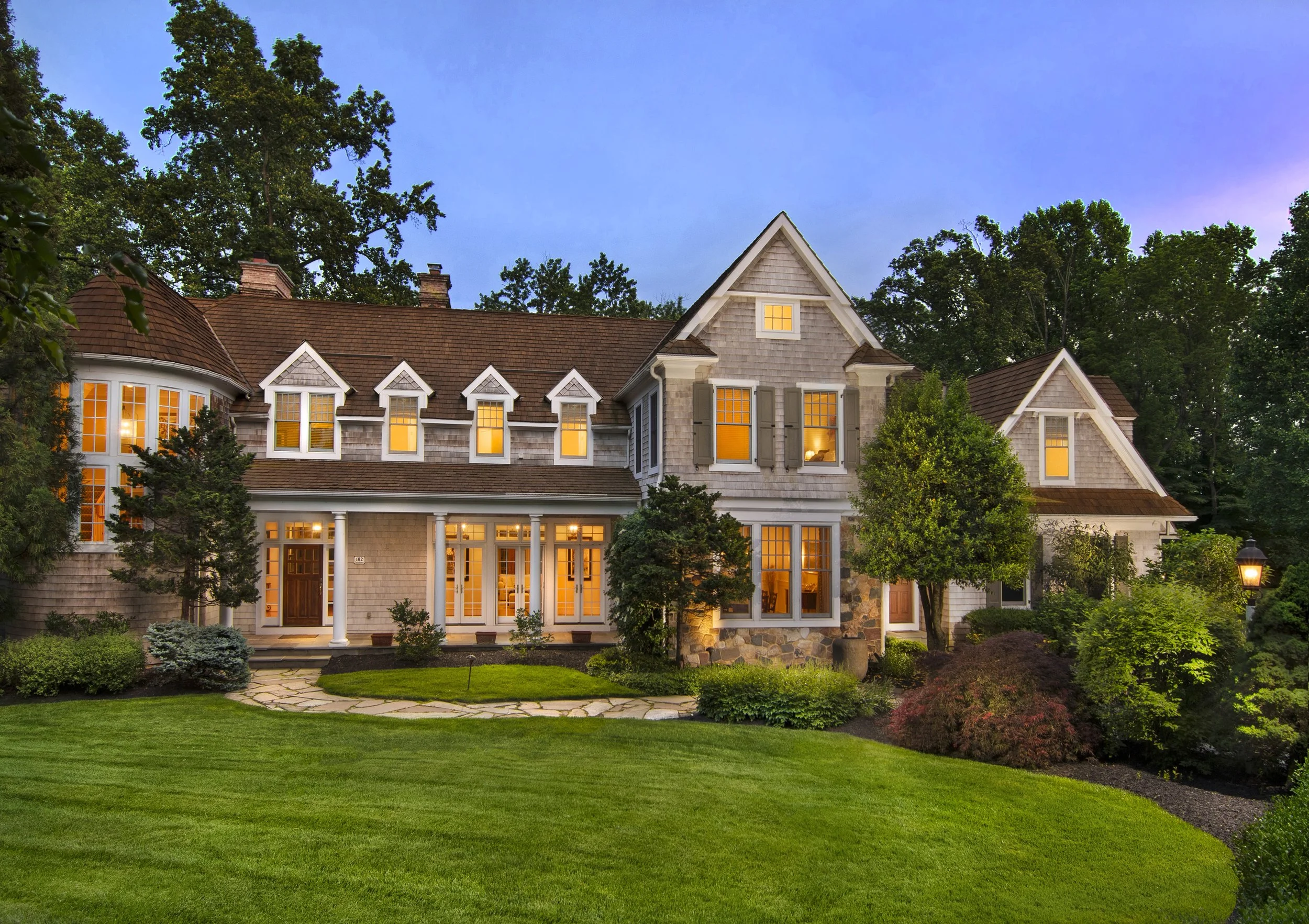 Twilight shot of a large, two-story house with turret and numerous windows illuminated from within, surrounded by a well-maintained lawn and lush trees.