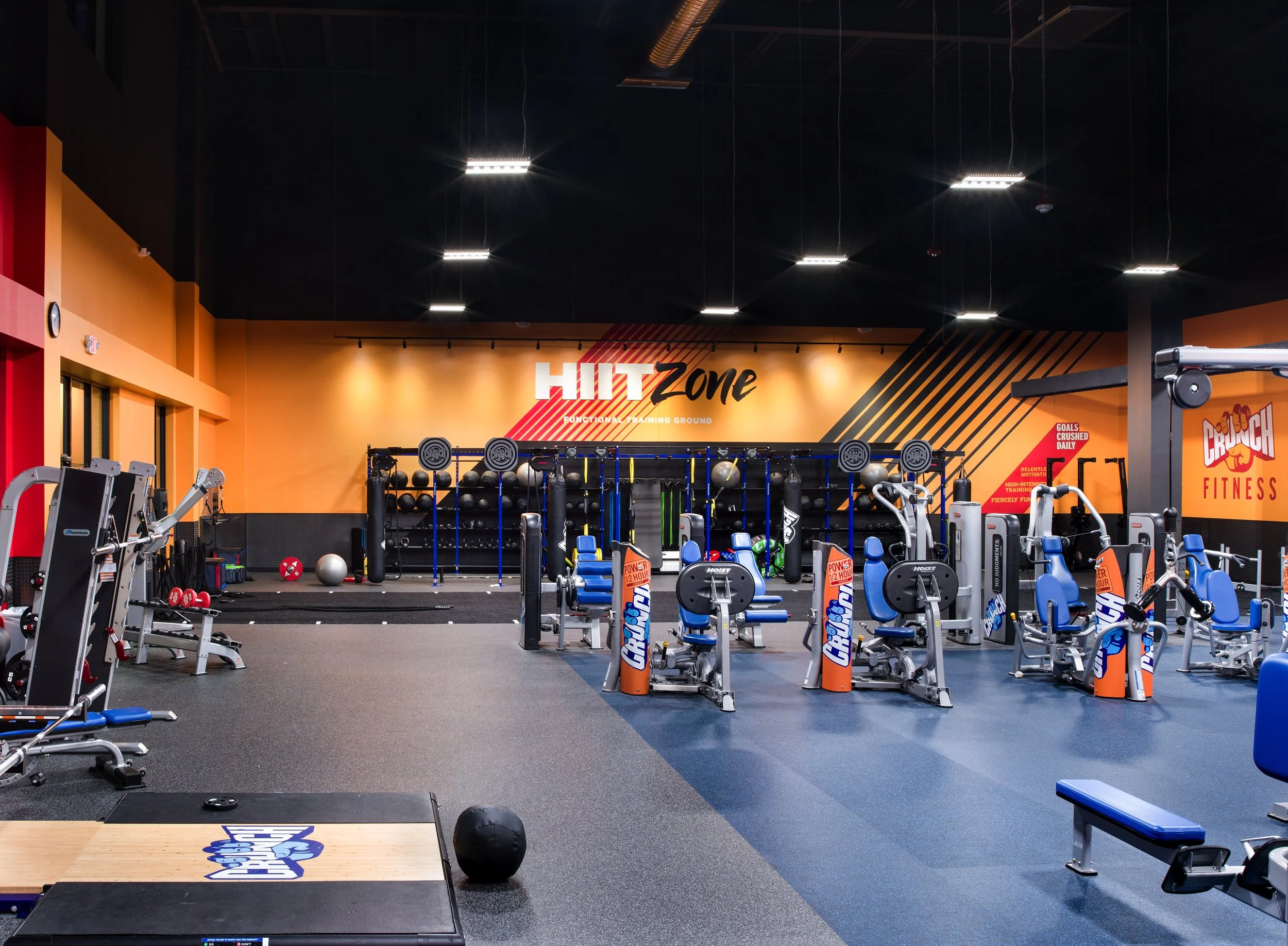 Inside a fitness gym with various workout equipment, including weight machines, medicine balls, and a pull-up station, with a large wall sign that reads 'HIIT Zone' and motivational posters.