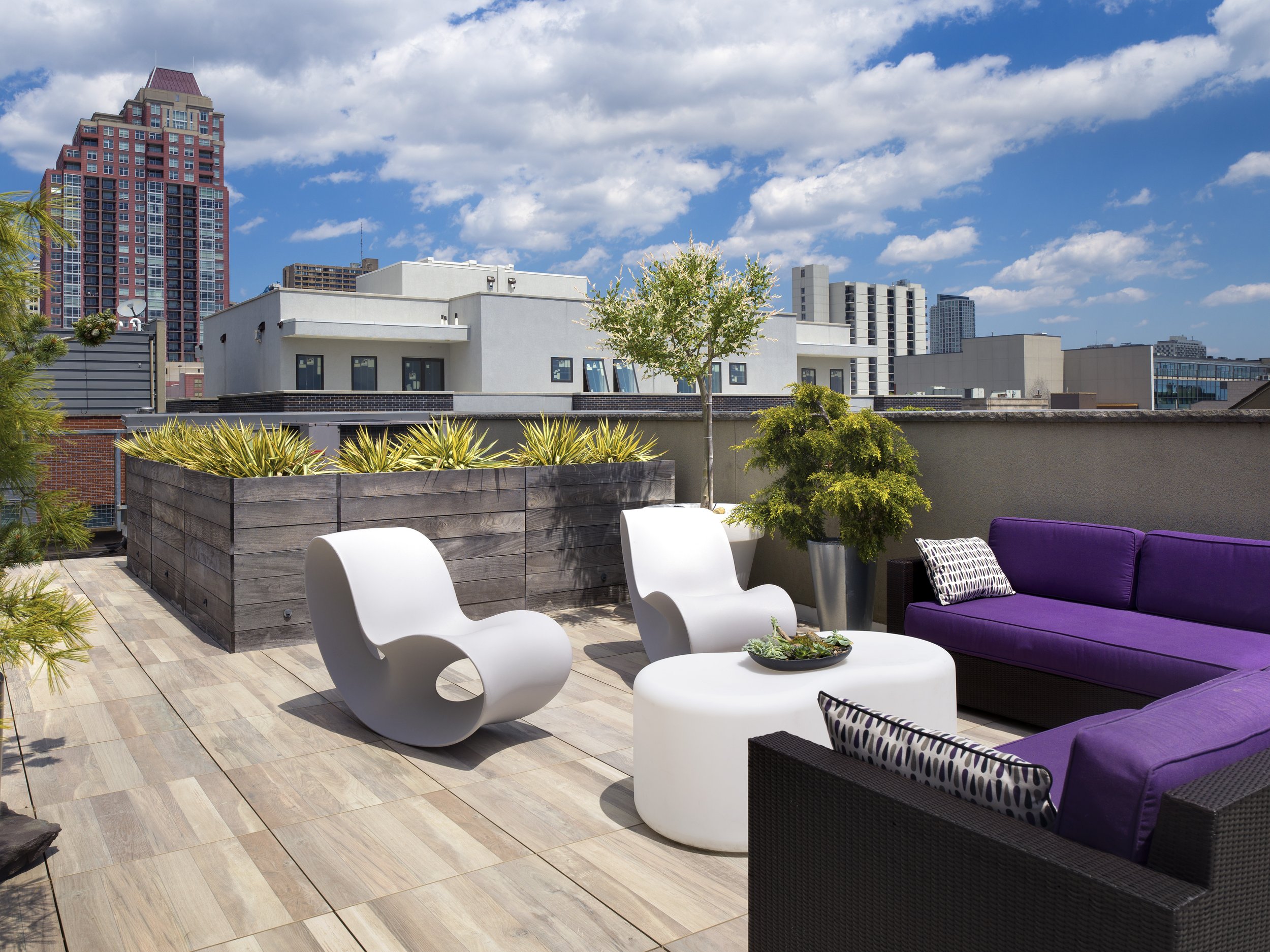 City skyline with modern buildings, a rooftop terrace with outdoor seating, potted plants, and a partly cloudy sky.