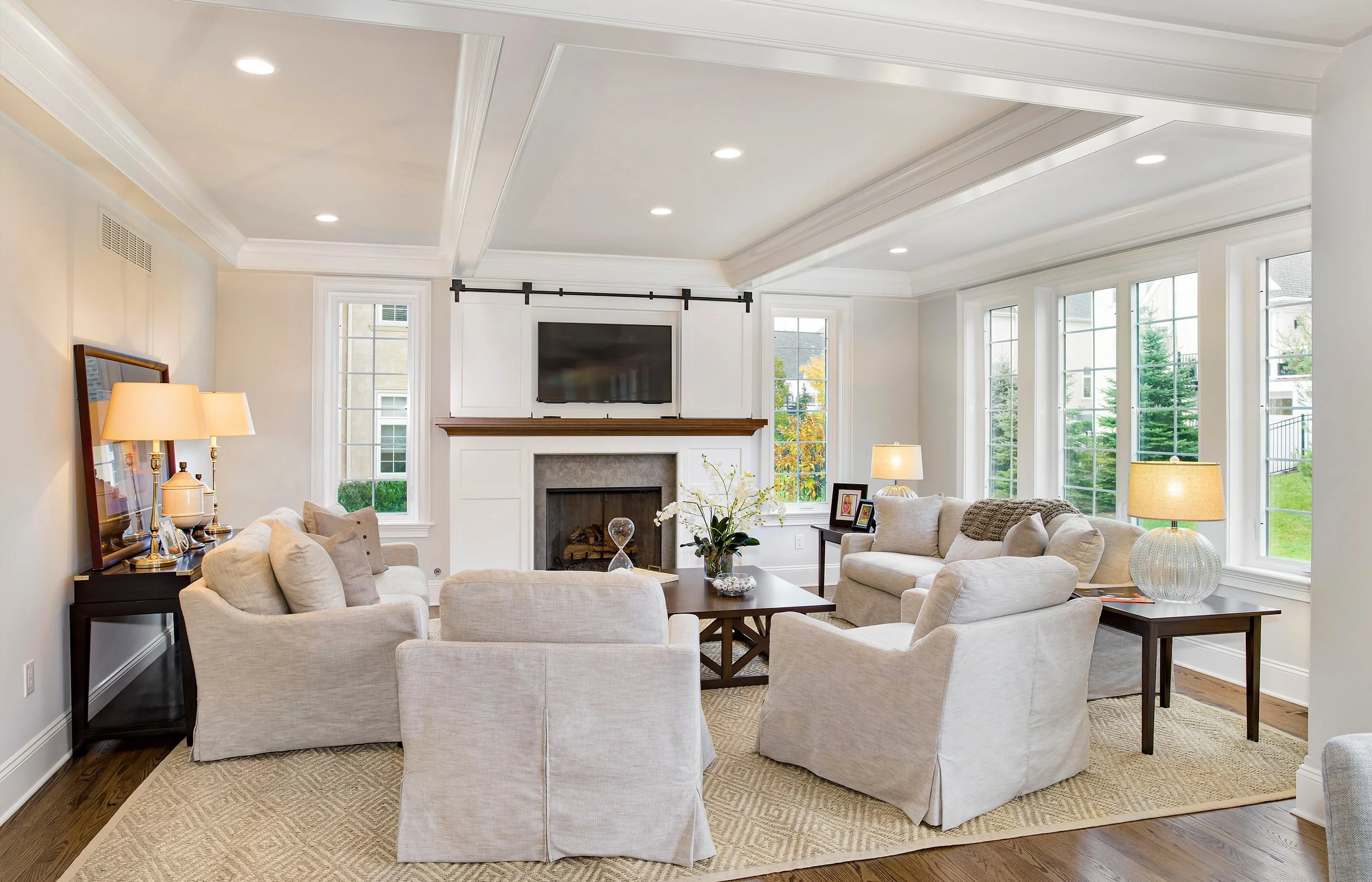 Bright living room with beige sofas, a fireplace, multiple windows, and a mounted TV above the fireplace.