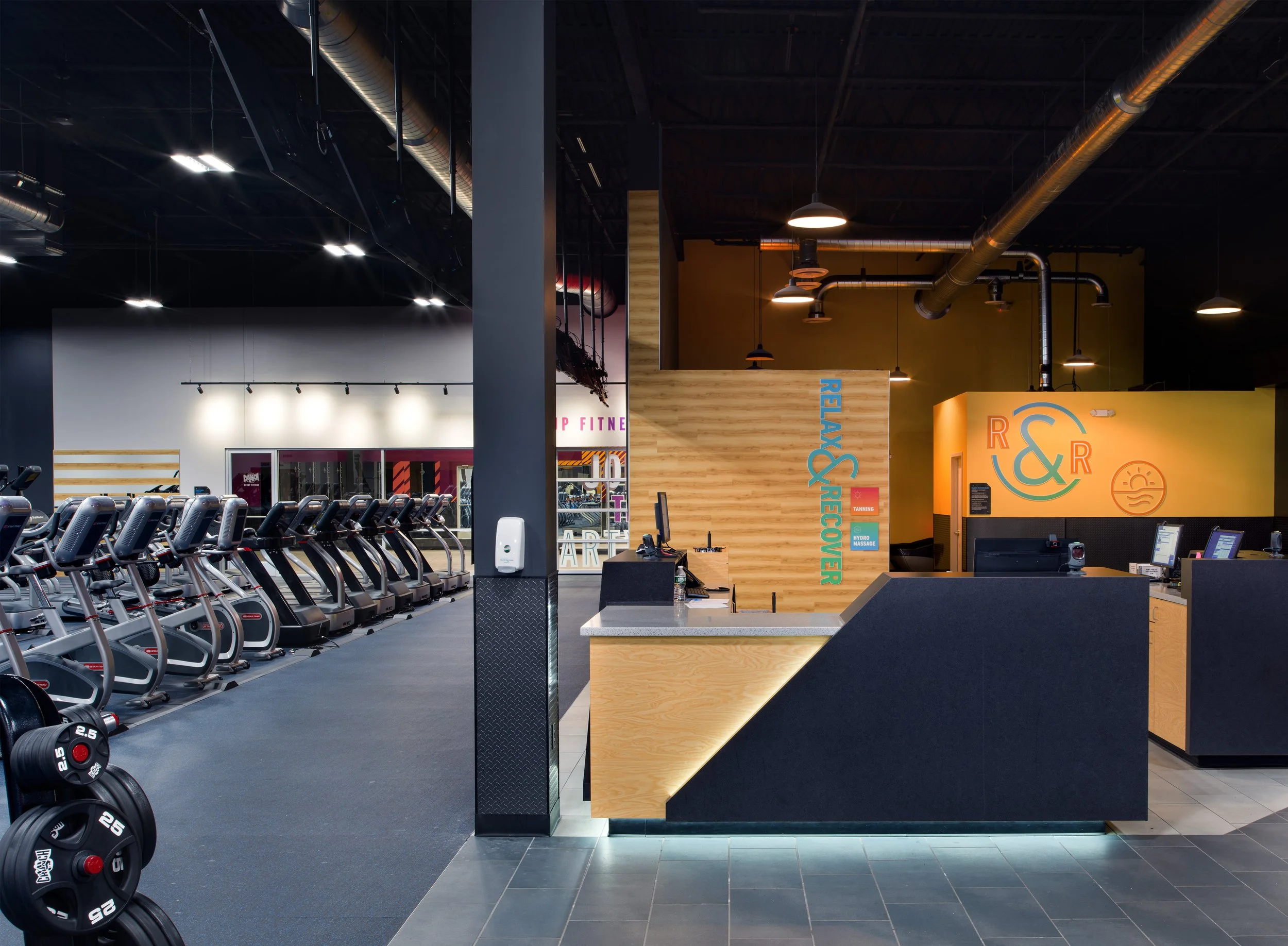 Interior of a gym with cardio machines in the foreground and a reception counter with wooden accents and colorful signage in the background, illuminated by ceiling lights and exposed ductwork.