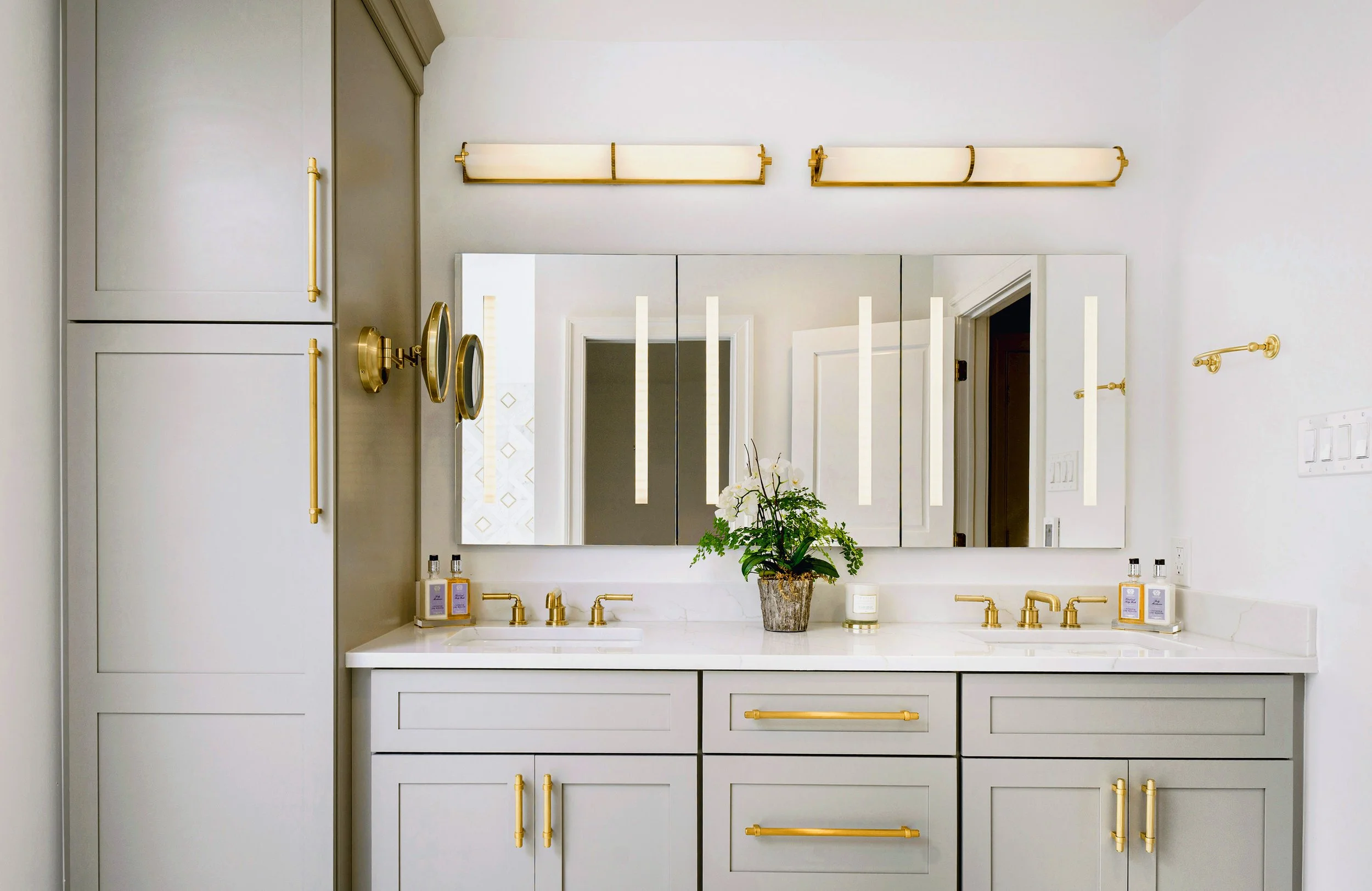 A modern bathroom vanity with white cabinets, gold hardware, and a white marble countertop. A large mirror with vertical light strips is above the sink, decorated with a potted orchid. Gold fixtures and accessories are on the countertop, with two bot