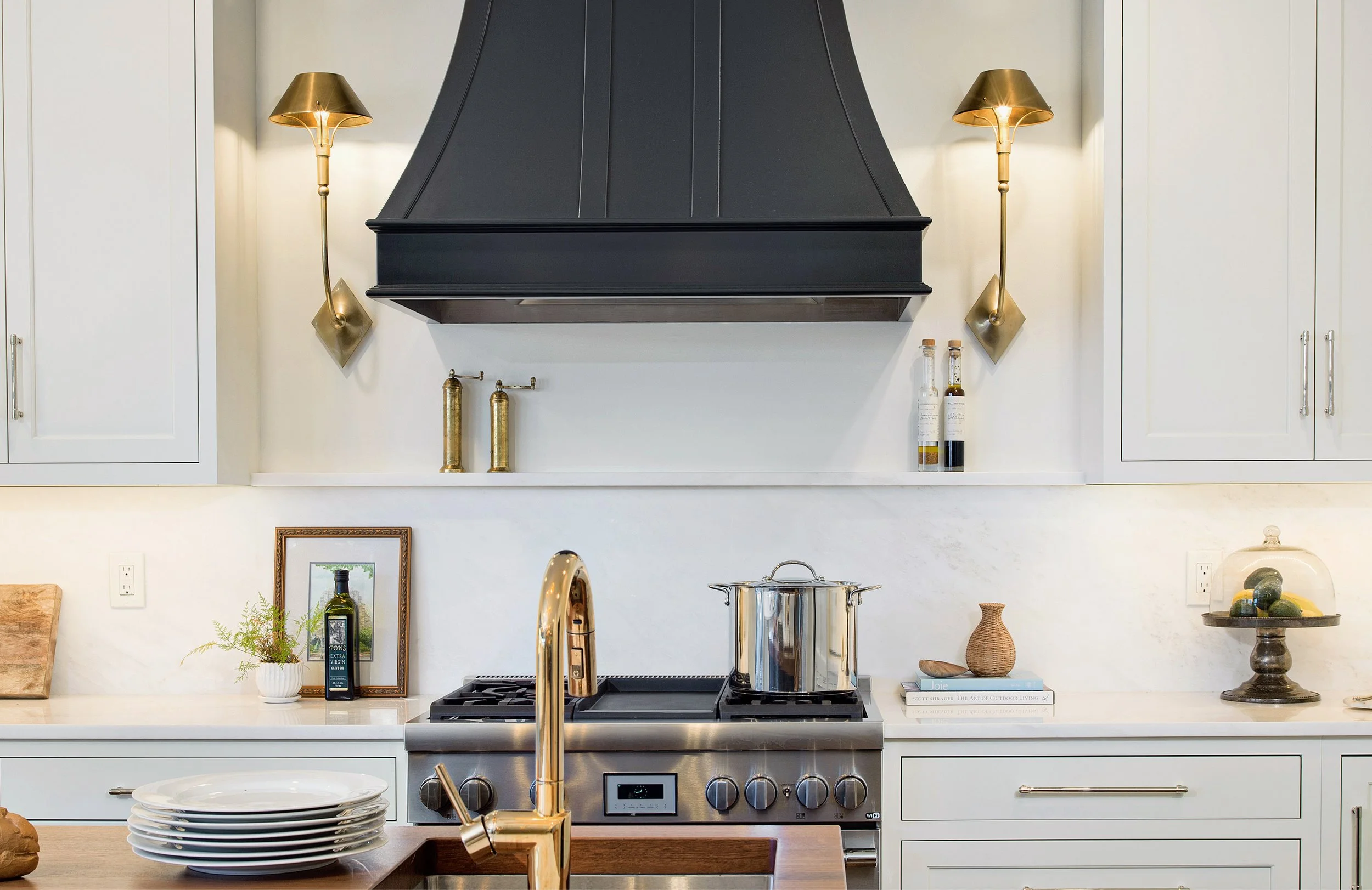 Modern kitchen with white cabinets, stainless steel stove, black range hood, gold wall sconces, and various decorative items and utensils on the countertop.