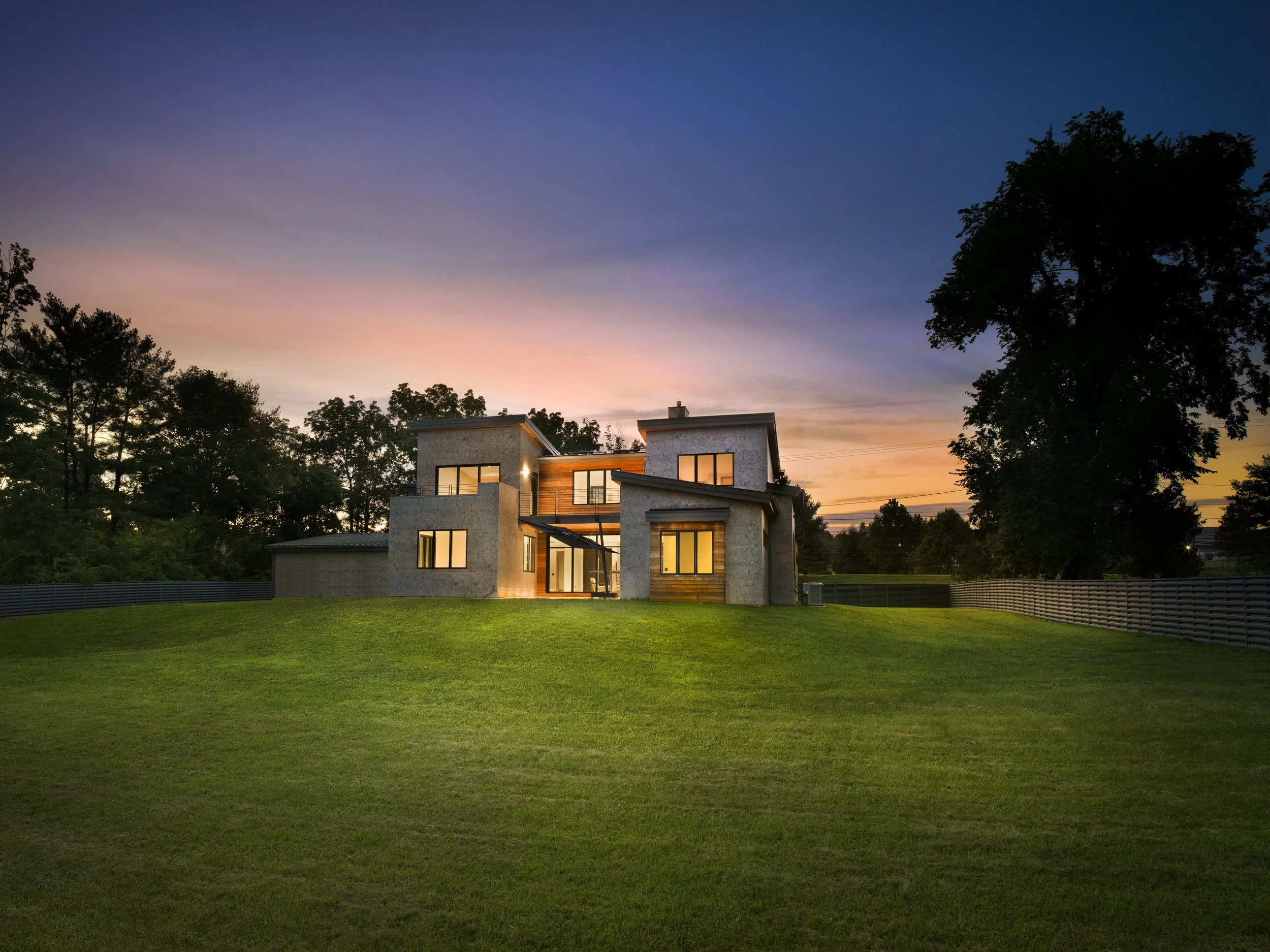 A modern house with large windows and a flat roof, illuminated from inside, set against a colorful sunset sky with pink, purple, and blue hues, surrounded by green grass and tall trees.