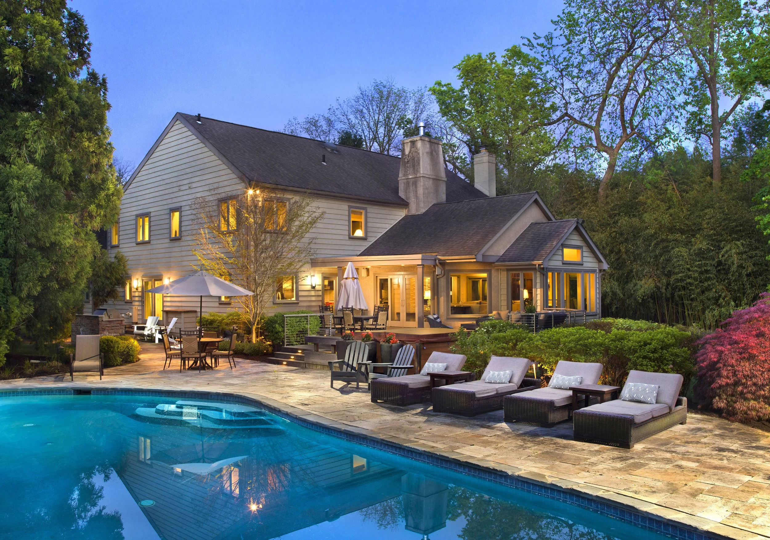 Twilight shot of a backyard with a swimming pool, outdoor lounge chairs, a dining area with a table and umbrella, and a large house with multiple windows and lighting at dusk.