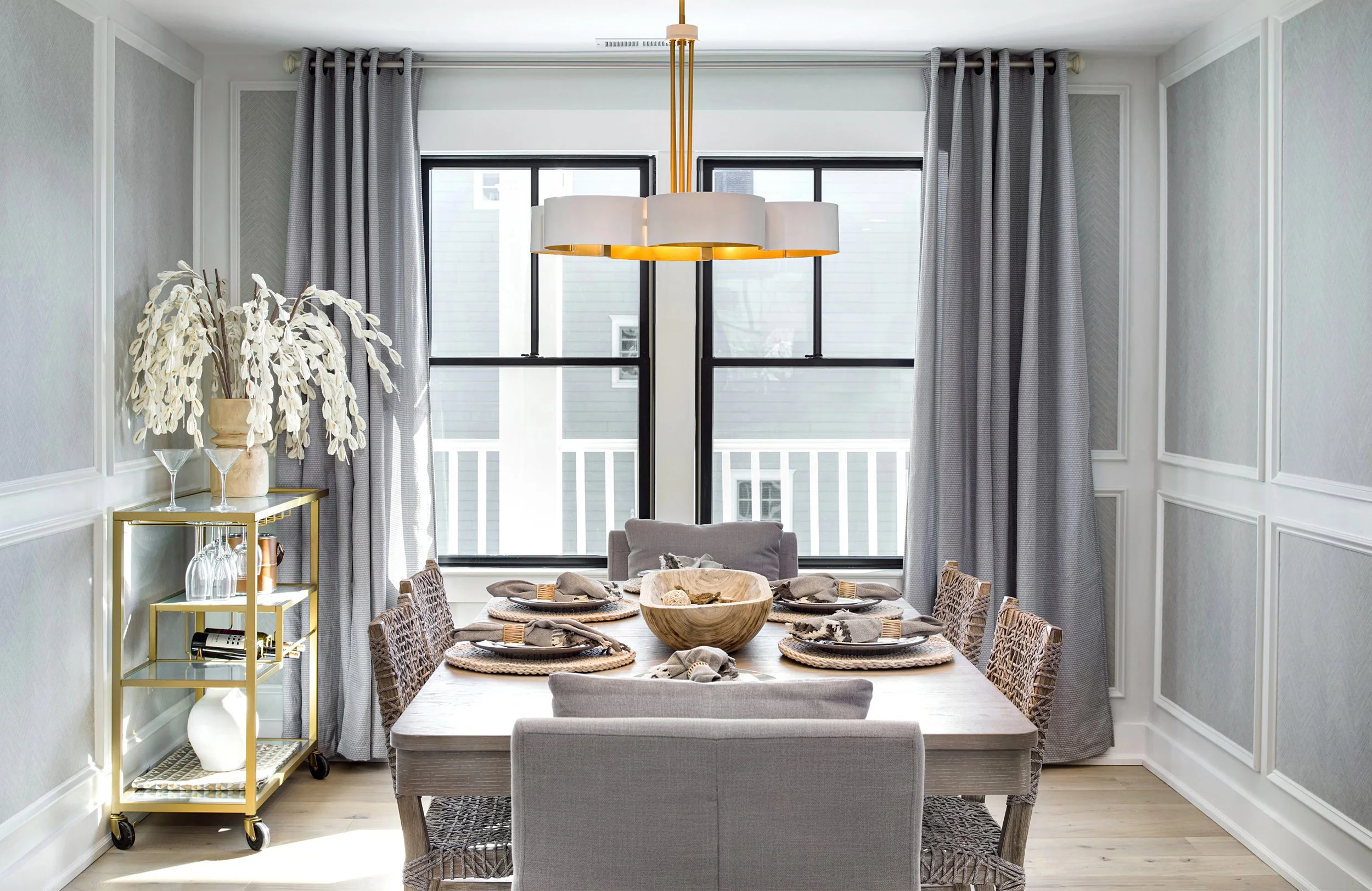 "Beachy" Dining room with a wooden table set with six place settings, a wooden bowl centerpiece, and upholstered chairs. Large windows with gray curtains, a gold bar cart with decorative items, and a modern white and gold chandelier overhead.