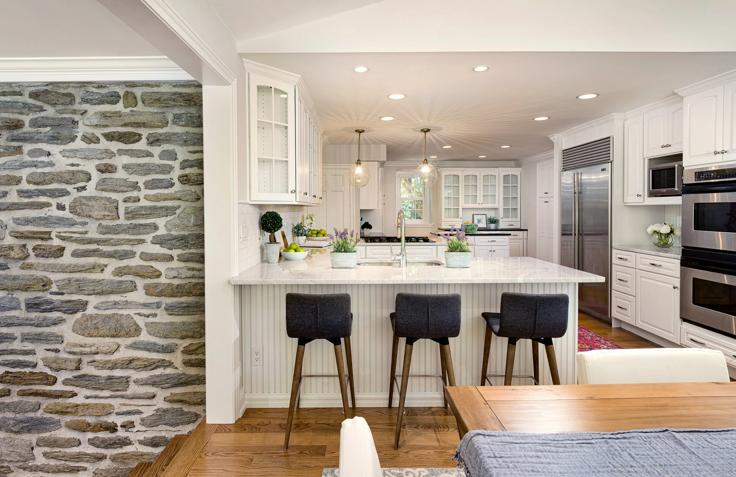 A modern kitchen with white cabinets, a marble island with three black chairs, stainless steel appliances, a stone accent wall, and various decorative plants and bowls on the countertop.
