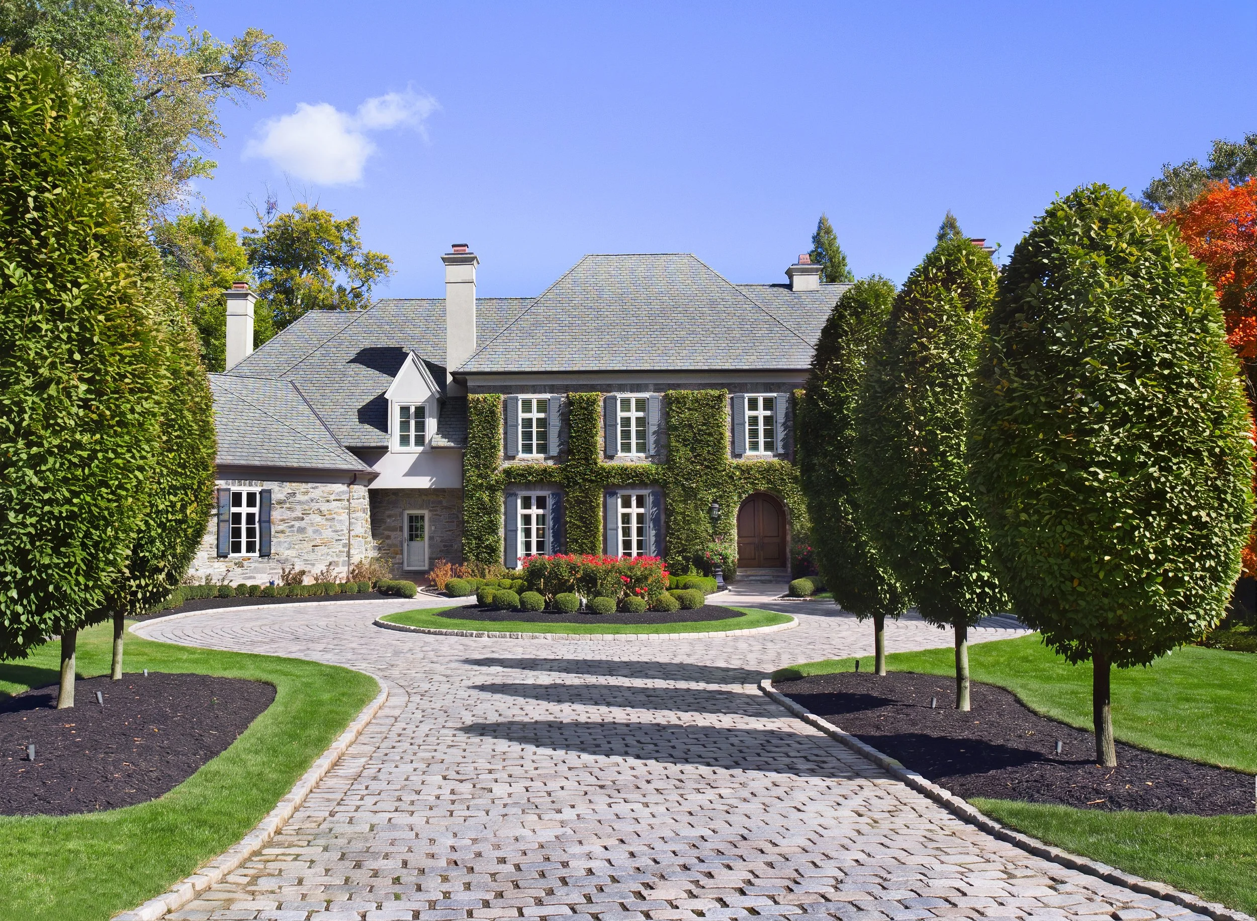 Elegant ivy-covered stone house with slate roof, manicured hedges, landscaped garden, and cobblestone driveway under a blue sky