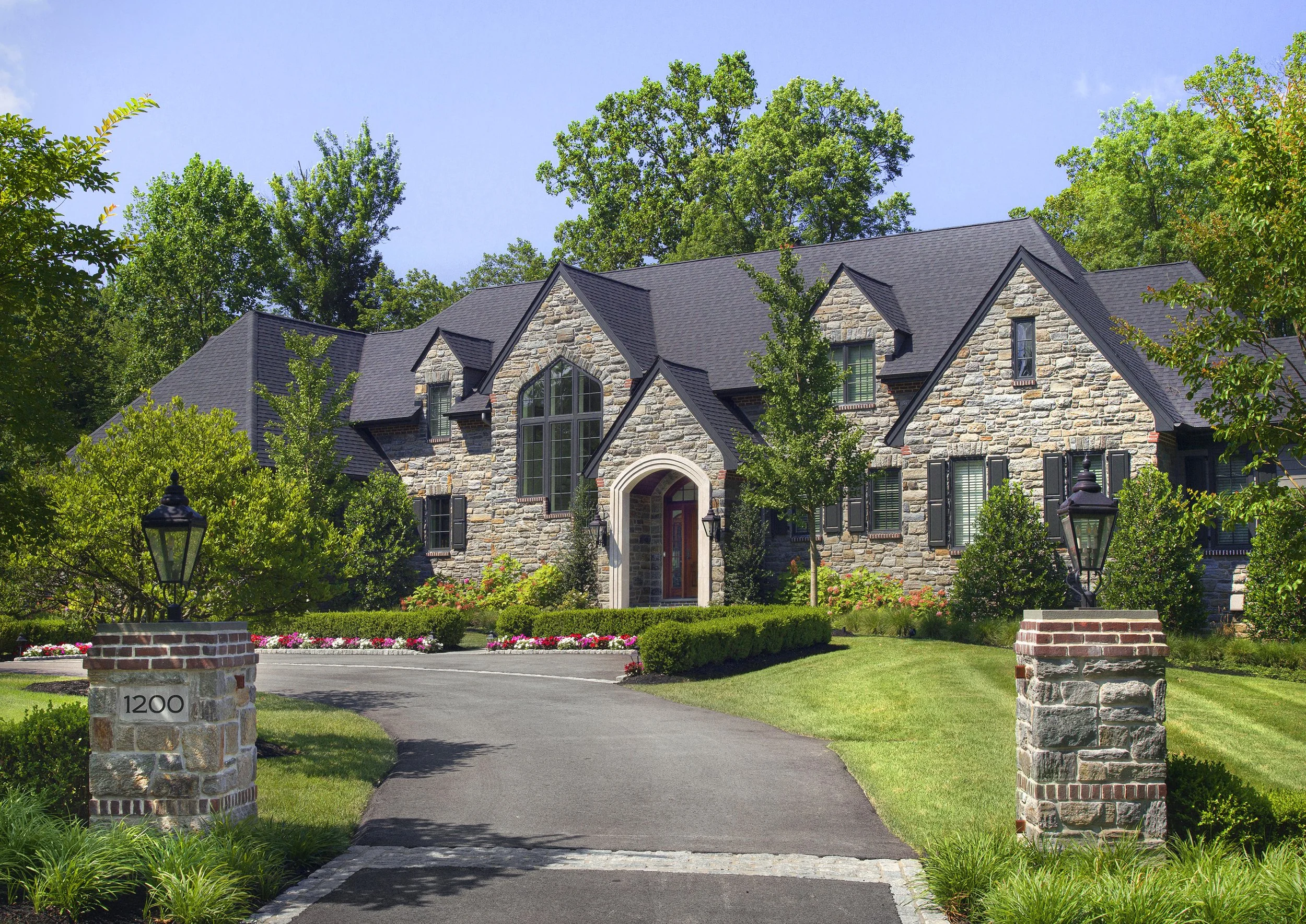 Large stone house with black roof, arched doorway, and multiple windows surrounded by lush green trees and landscaping. Brick pillars with lamps line the driveway, and the house number is 1200.