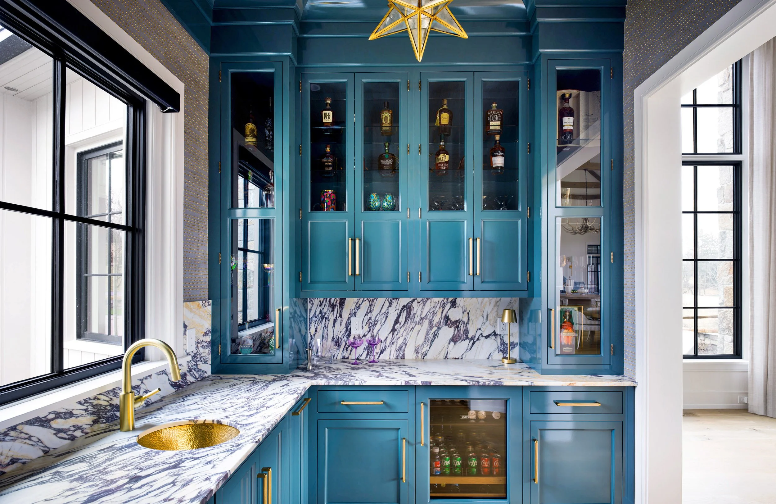 View of a blue kitchen with glass-front cabinets, marble countertops, a gold sink, and windows providing natural light.