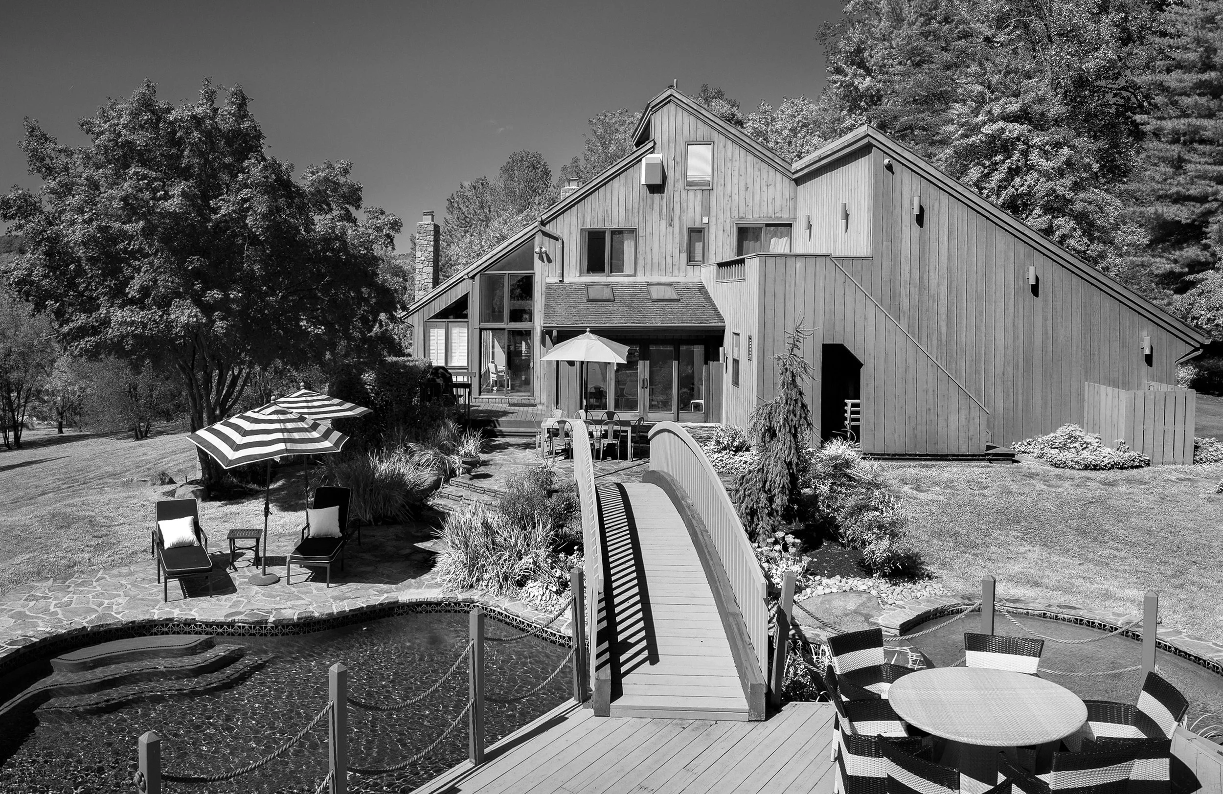 Black and white photo of a modern house with a sloped roof, surrounded by trees. In the foreground, there is a patio with lounge chairs, a table with an umbrella, and a small pond with a bridge crossing over it.