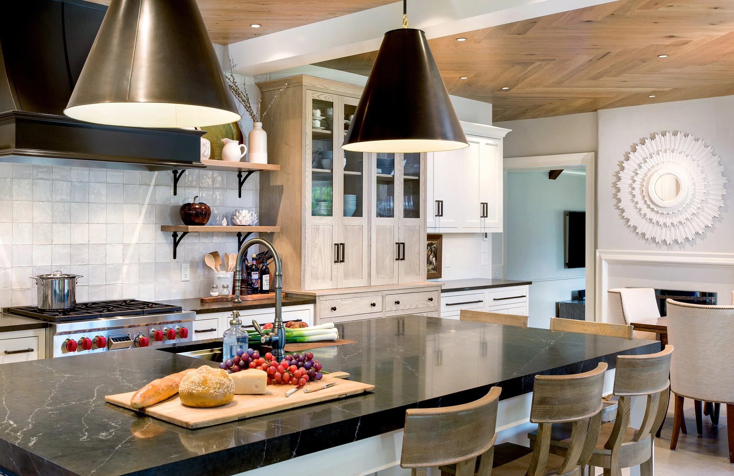 Modern kitchen with black marble island, hanging black pendant lights, white cabinets, and open shelving with dishes and decorative items. There are fresh fruits and bread on the island, and a view into the adjacent dining and living area.