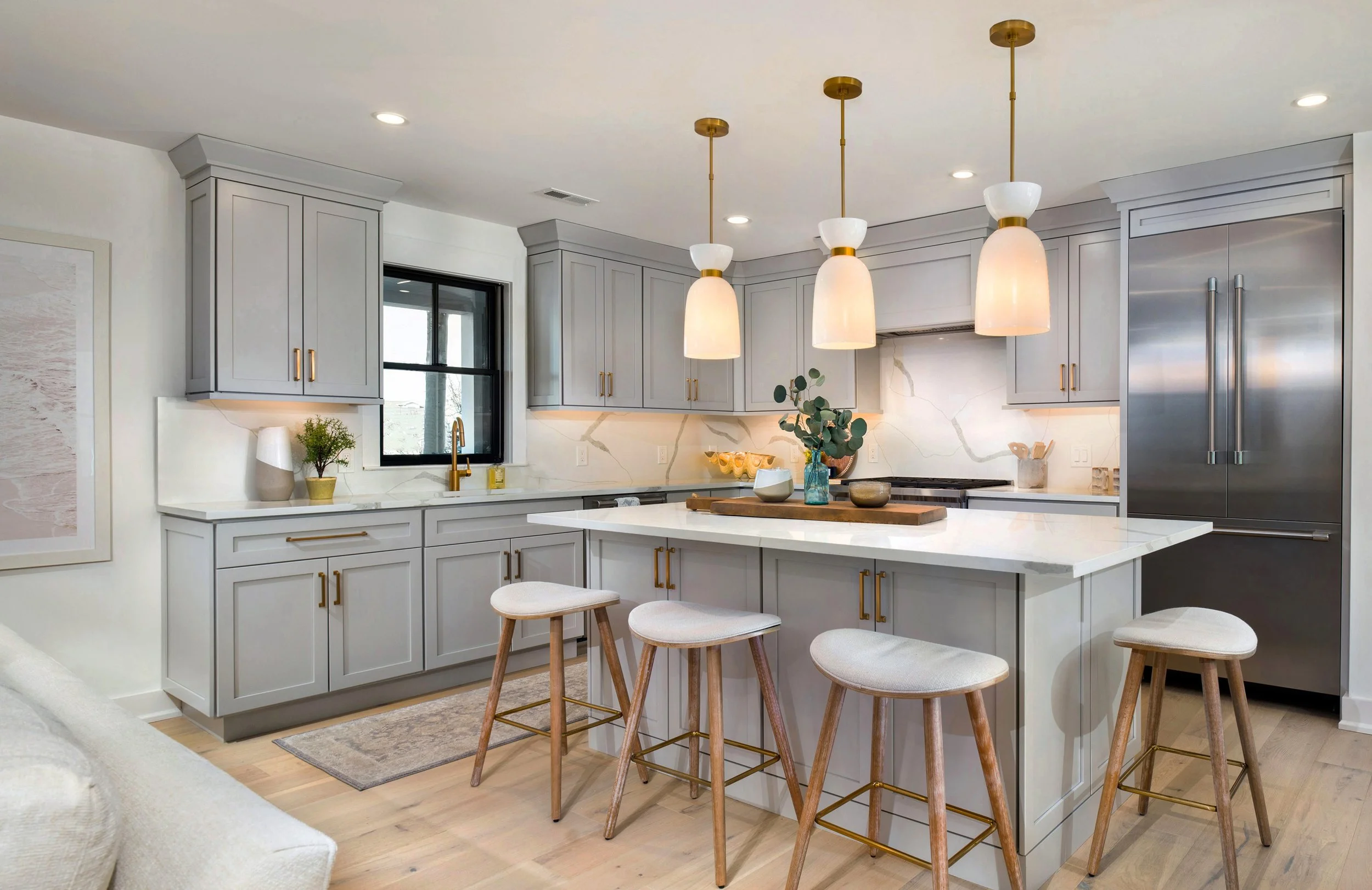 Modern kitchen with white cabinets, marble backsplash, stainless steel refrigerator, kitchen island with four stools, pendant lights, and decorative plants.