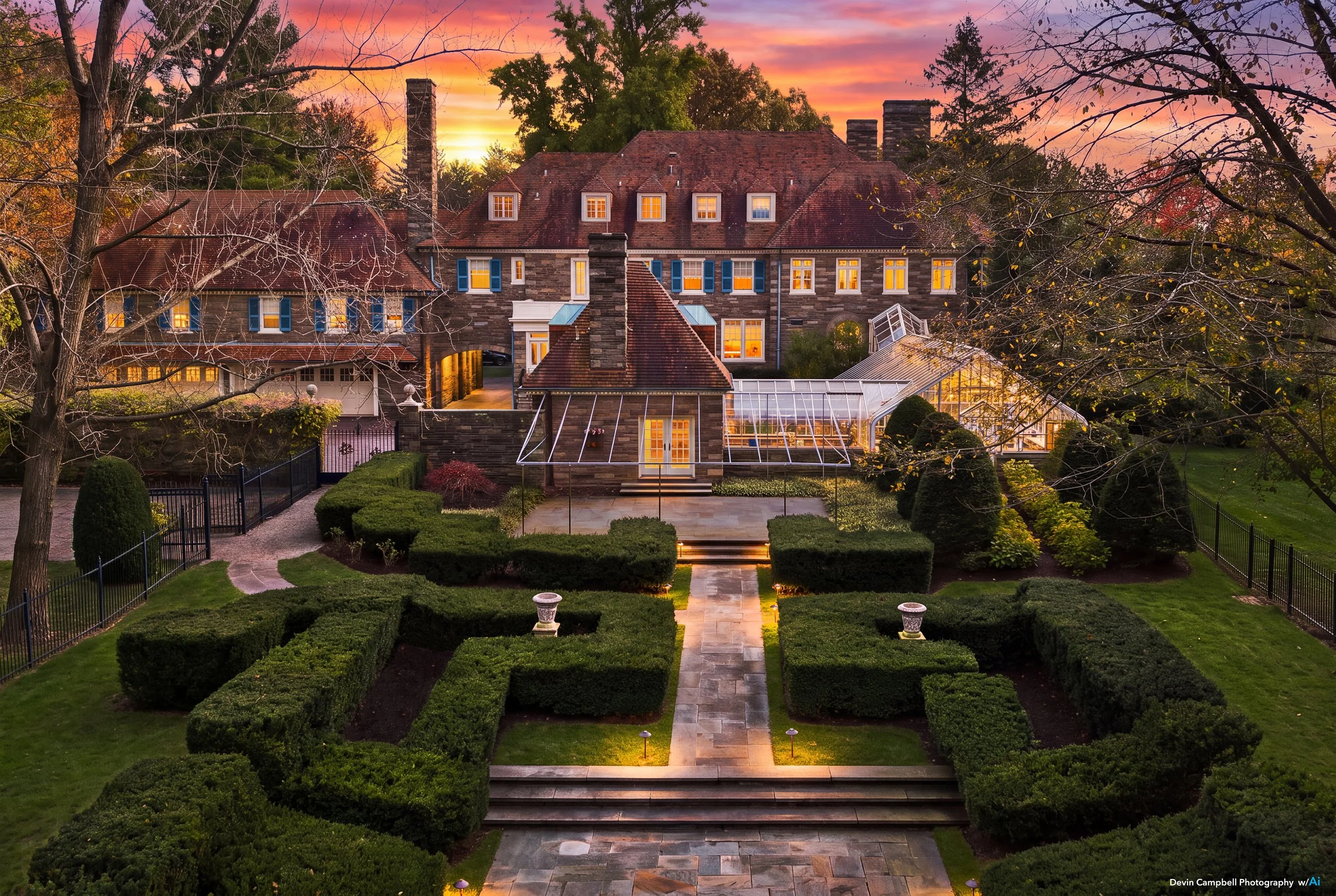 Historic stone mansion with glowing windows, greenhouse, and formal hedge garden at sunset in Fall
