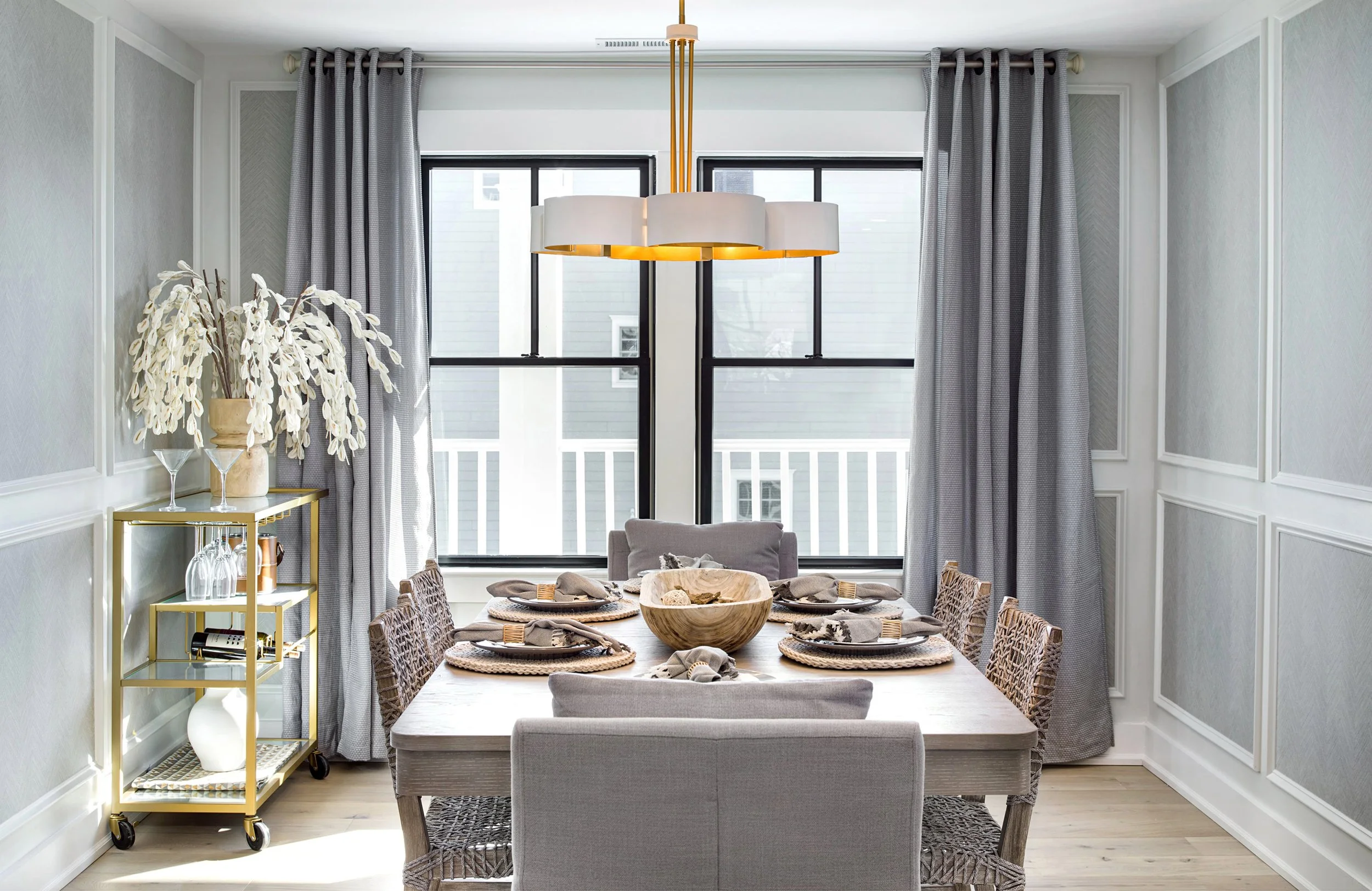 "Beachy" Dining room with a wooden table set with six place settings, a wooden bowl centerpiece, and upholstered chairs. Large windows with gray curtains, a gold bar cart with decorative items, and a modern white and gold chandelier overhead.