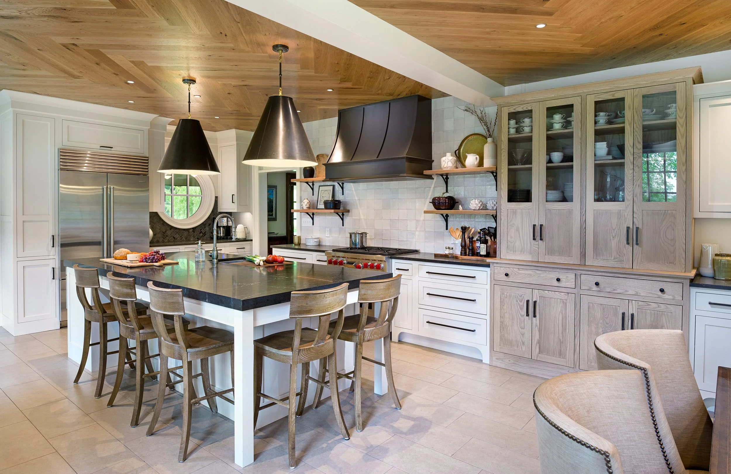 A modern kitchen with a large island, white cabinetry, open shelving, and stainless steel appliances, including a double-door refrigerator, custom range hood all illuminated by pendant lights and natural light from a round window.