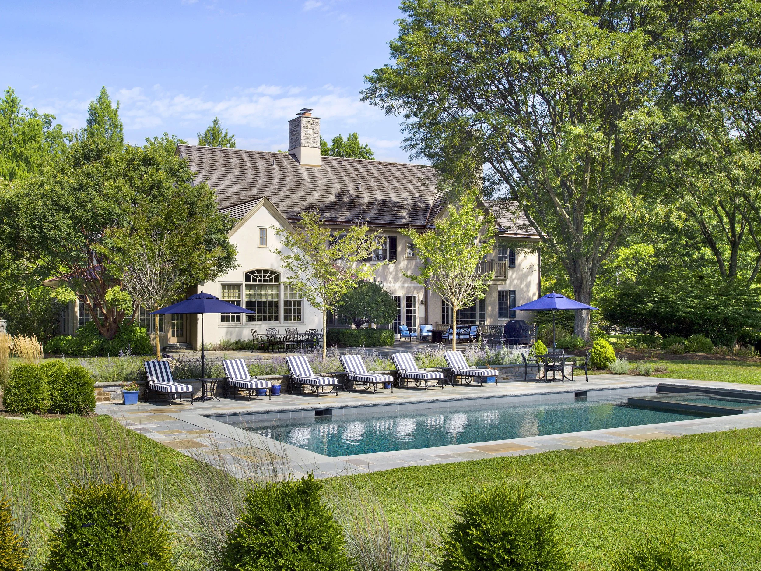 Backyard view of a house with a swimming pool in the foreground, surrounded by greenery, trees, and outdoor furniture with umbrellas under a blue sky.