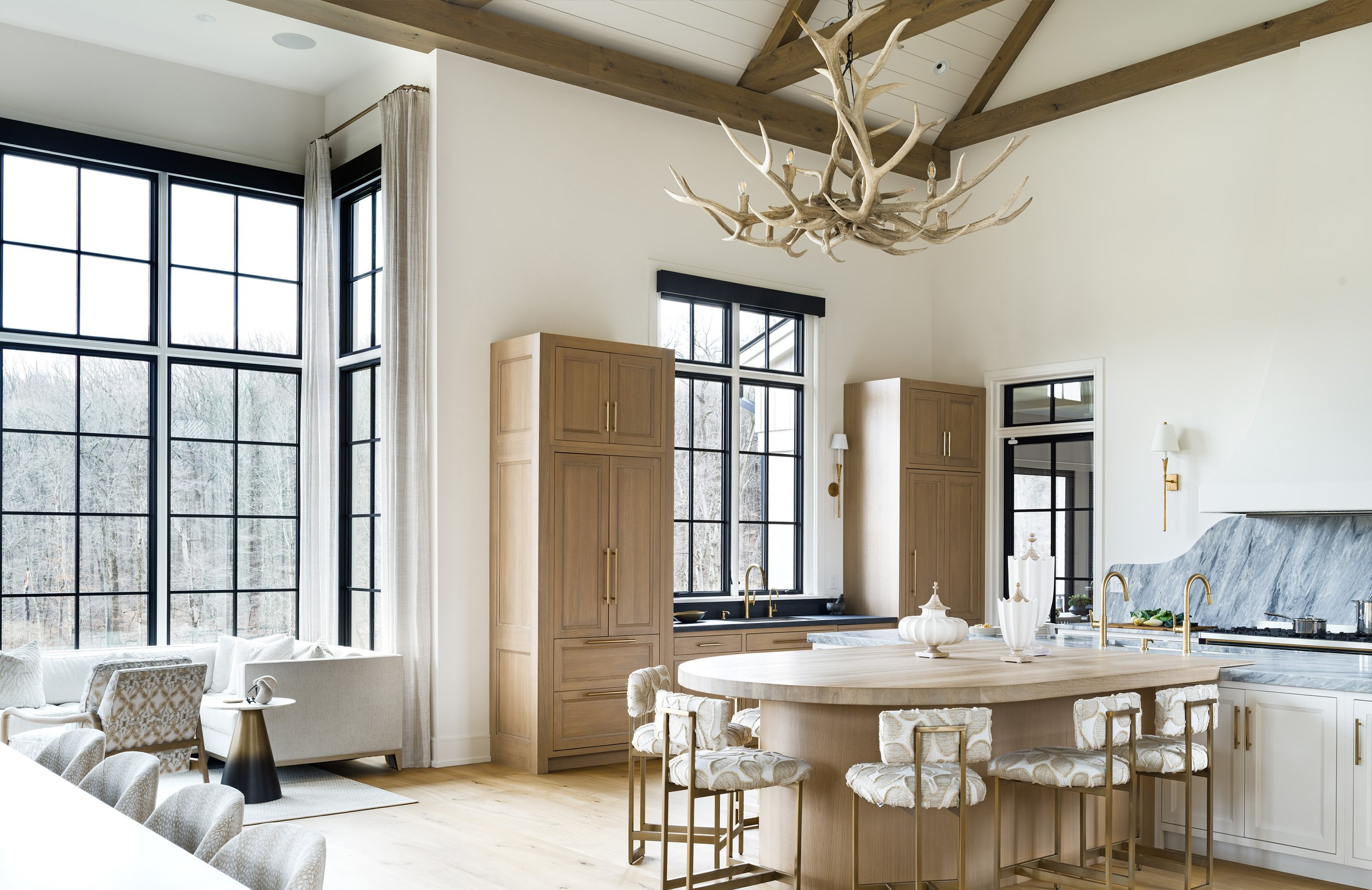 Bright luxury kitchen and living room with wood cabinetry, marble island, black-framed windows, exposed beams, and antler chandelier