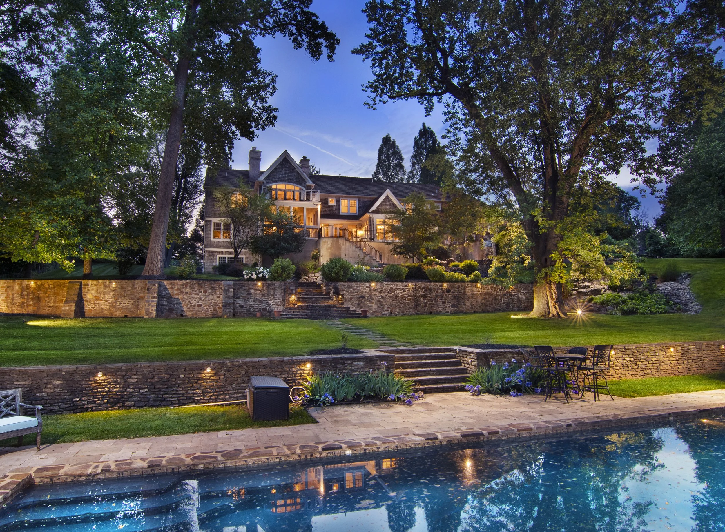 Twilight shot of a beautifully lit backyard at dusk with a swimming pool in the foreground, a stone patio with outdoor furniture, a well-maintained lawn, stone stairs leading up to a large house surrounded by trees and landscaped gardens.
