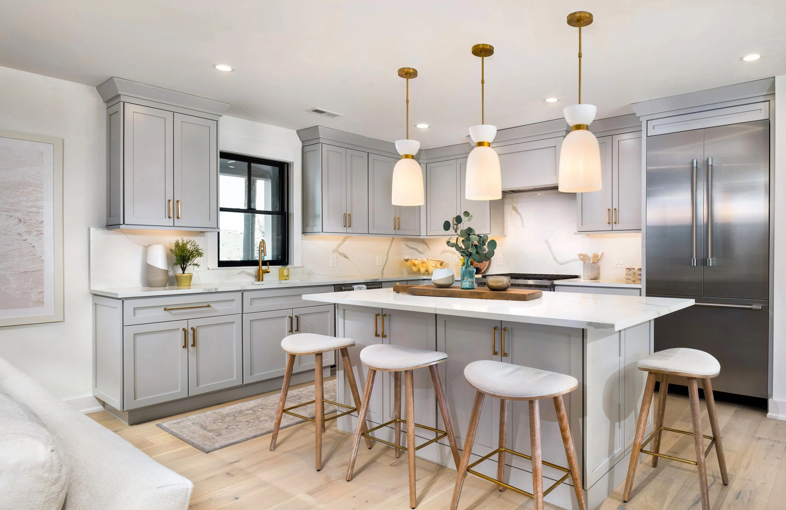 Modern kitchen with white cabinets, marble backsplash, stainless steel refrigerator, kitchen island with four stools, pendant lights, and decorative plants.