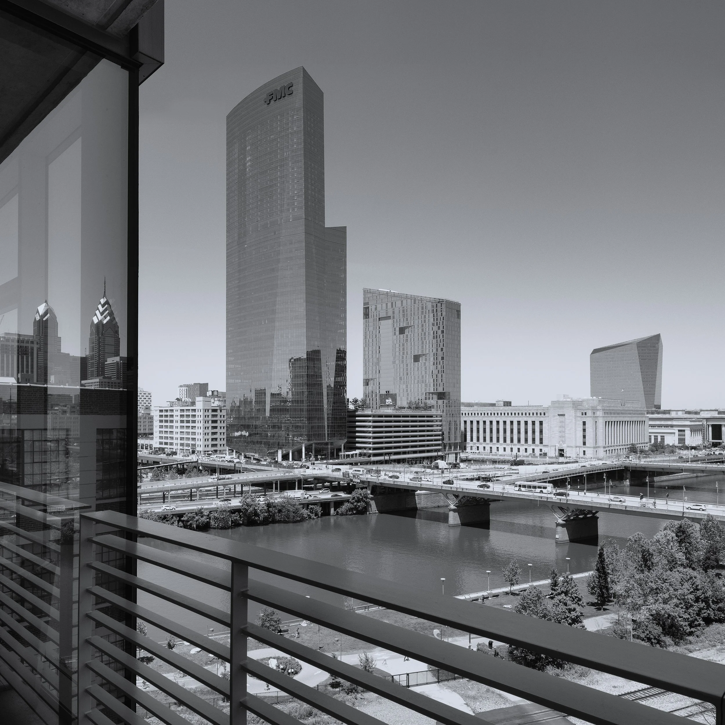 Black-and-white Philadelphia skyline viewed from a balcony, featuring the FMC Tower, Schuylkill River, bridges, and Center City buildings.