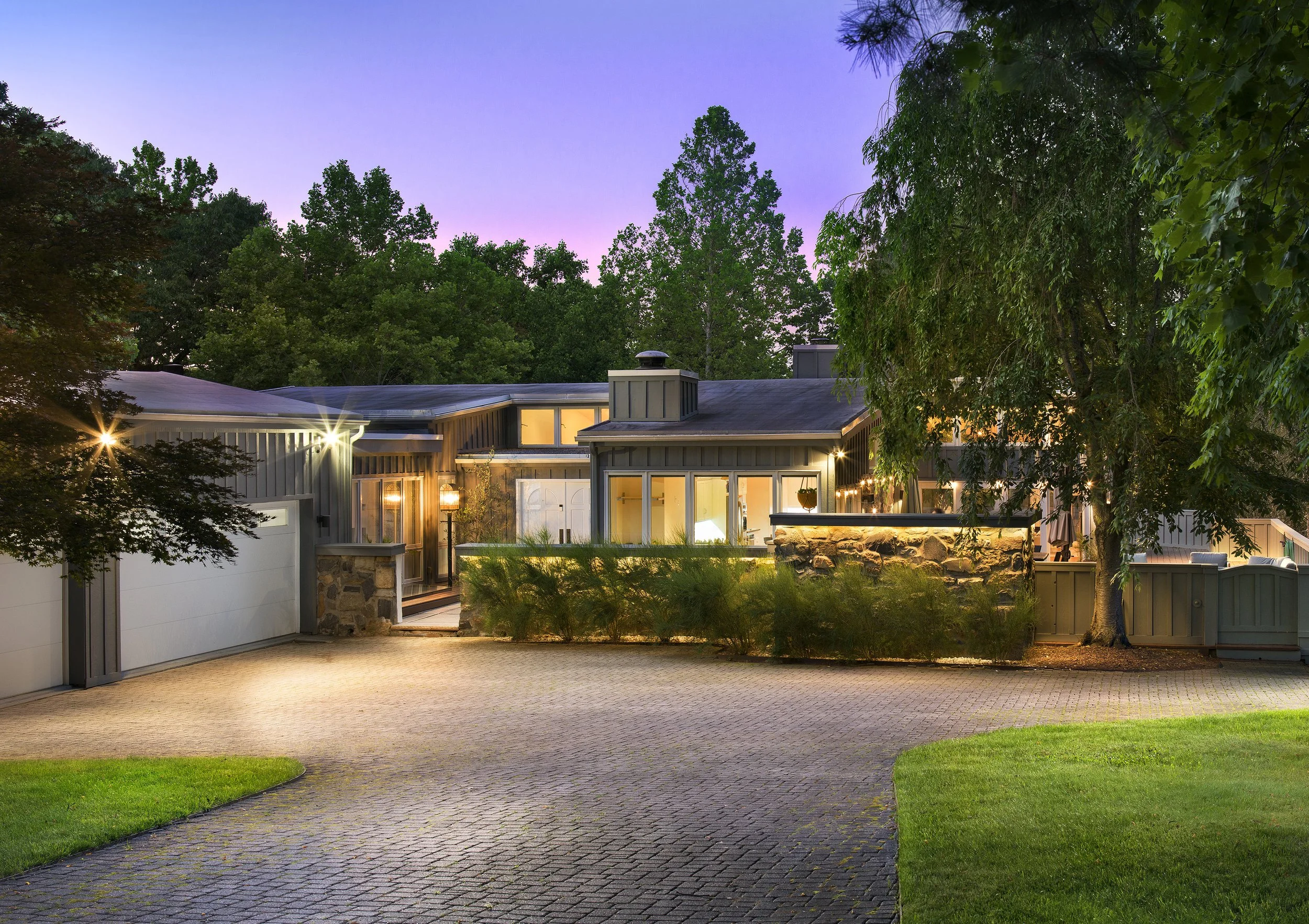 A modern house with large glass windows and doors, surrounded by trees, illuminated from inside, at dusk. The driveway is made of interlocking bricks, with well-kept grass on the sides.