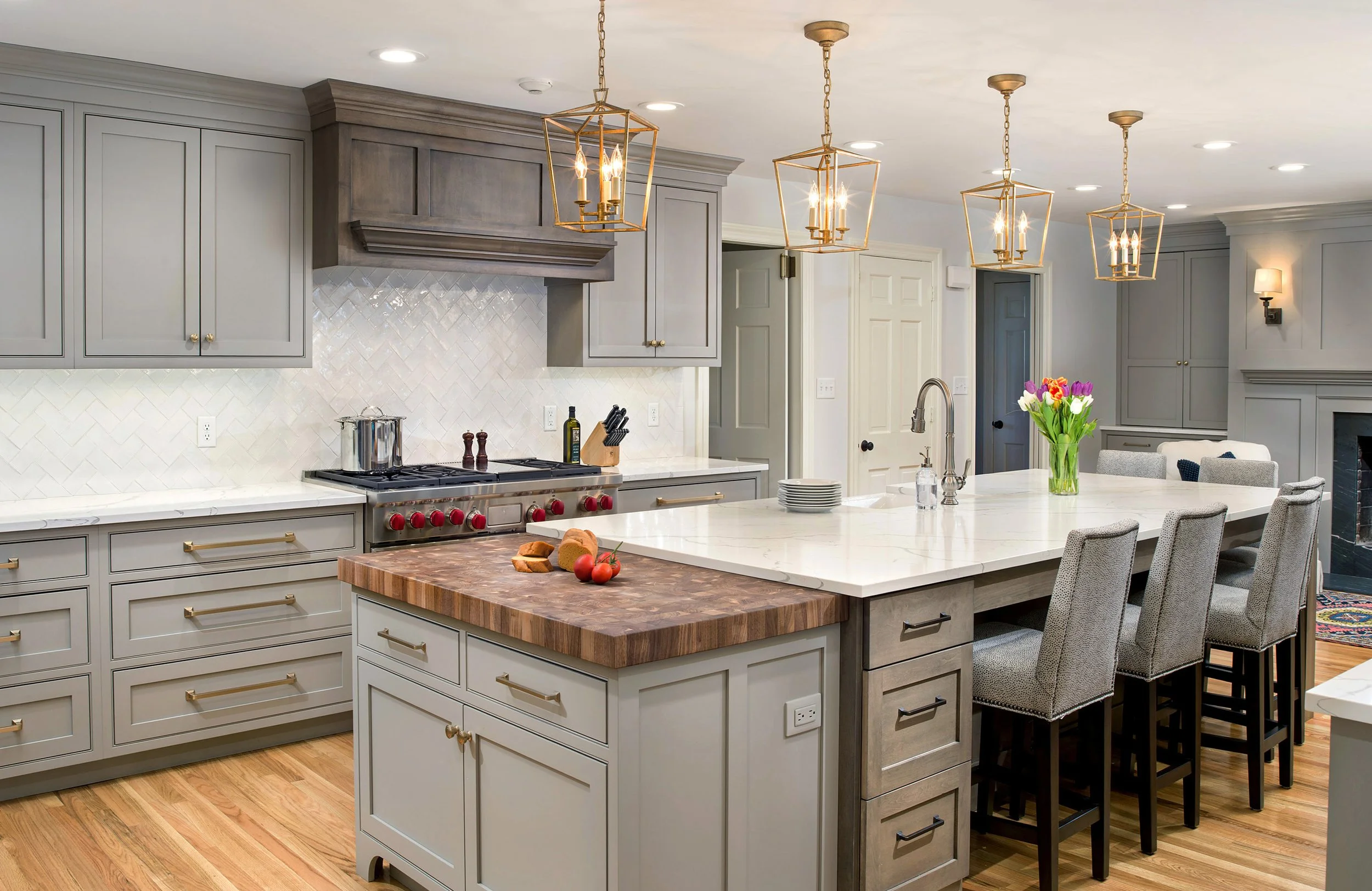 Modern kitchen with white cabinets, a large island with a white marble countertop, four upholstered bar stools, pendant lighting, a flower arrangement, and a gas stove with a wooden cutting board and fresh tomatoes.