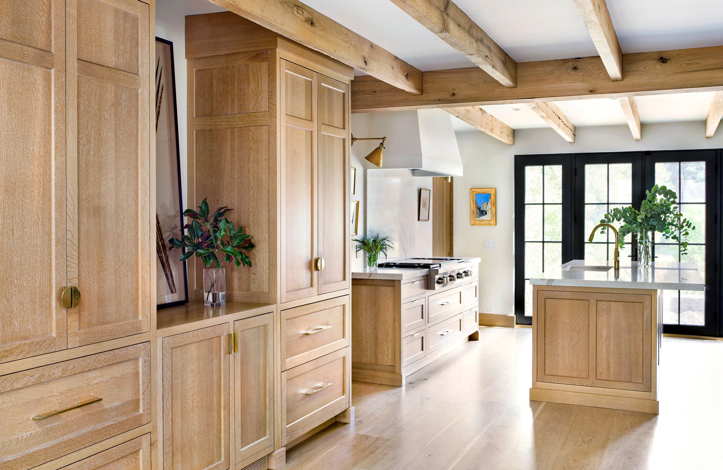 Modern kitchen with wooden cabinetry, a kitchen island with plants, black-framed glass doors, and exposed wooden ceiling beams.