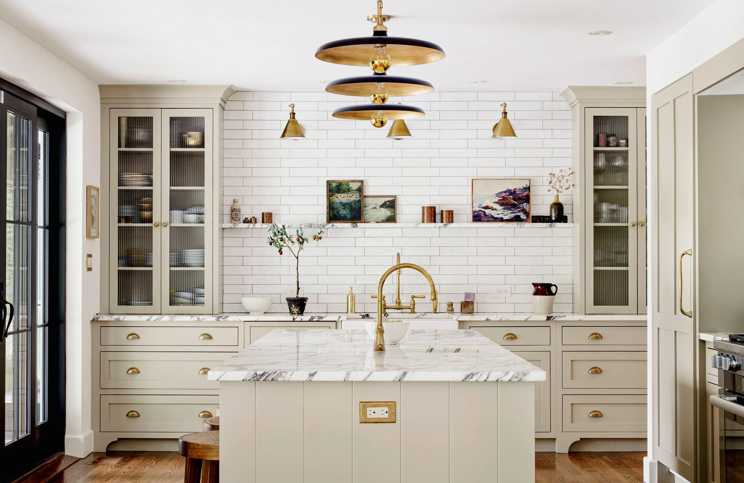 A modern kitchen with a white marble island, gold fixtures, and white cabinets with gold handles. There is a white brick backsplash and decorative paintings on a shelf. Gold and black pendant lights hang from the ceiling, and there are glass-front ca