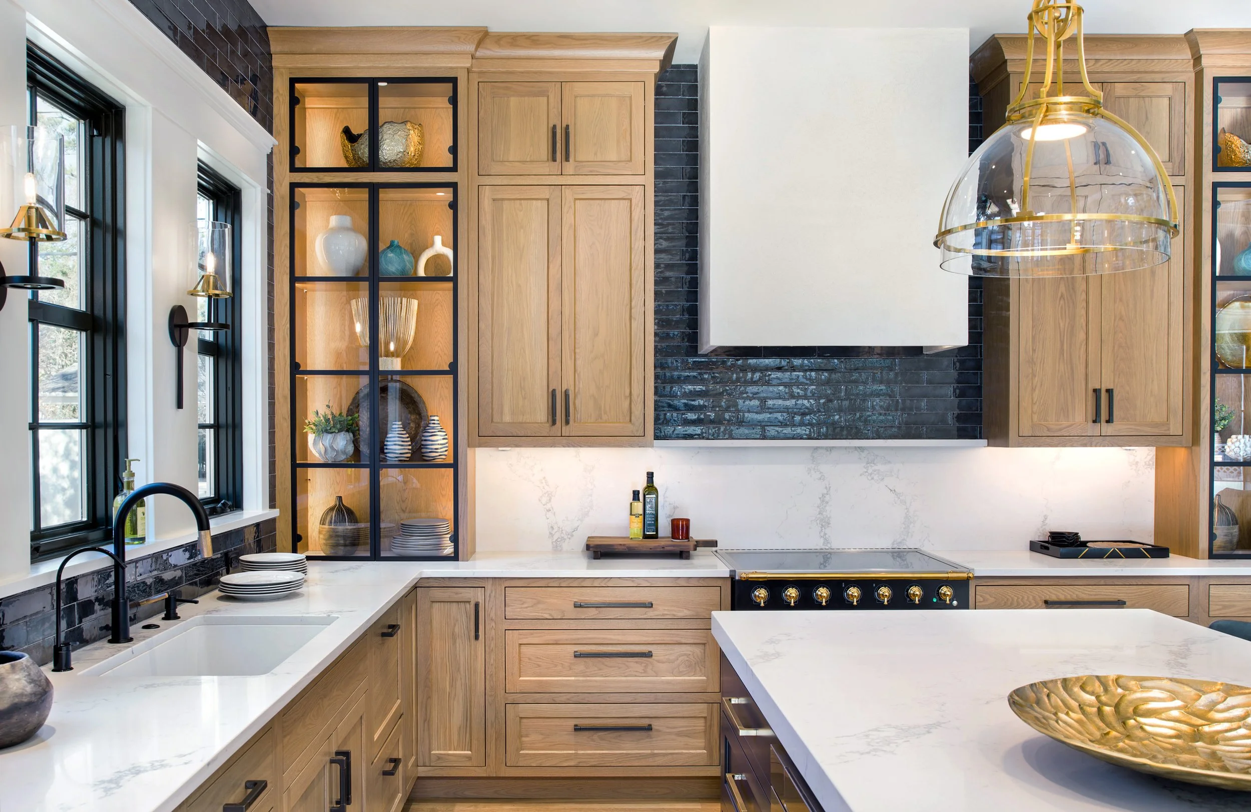 Modern kitchen with light wooden cabinets, white marble countertops, black window frames, black backsplash, and gold accents in light fixture and decor. Black subway tile.