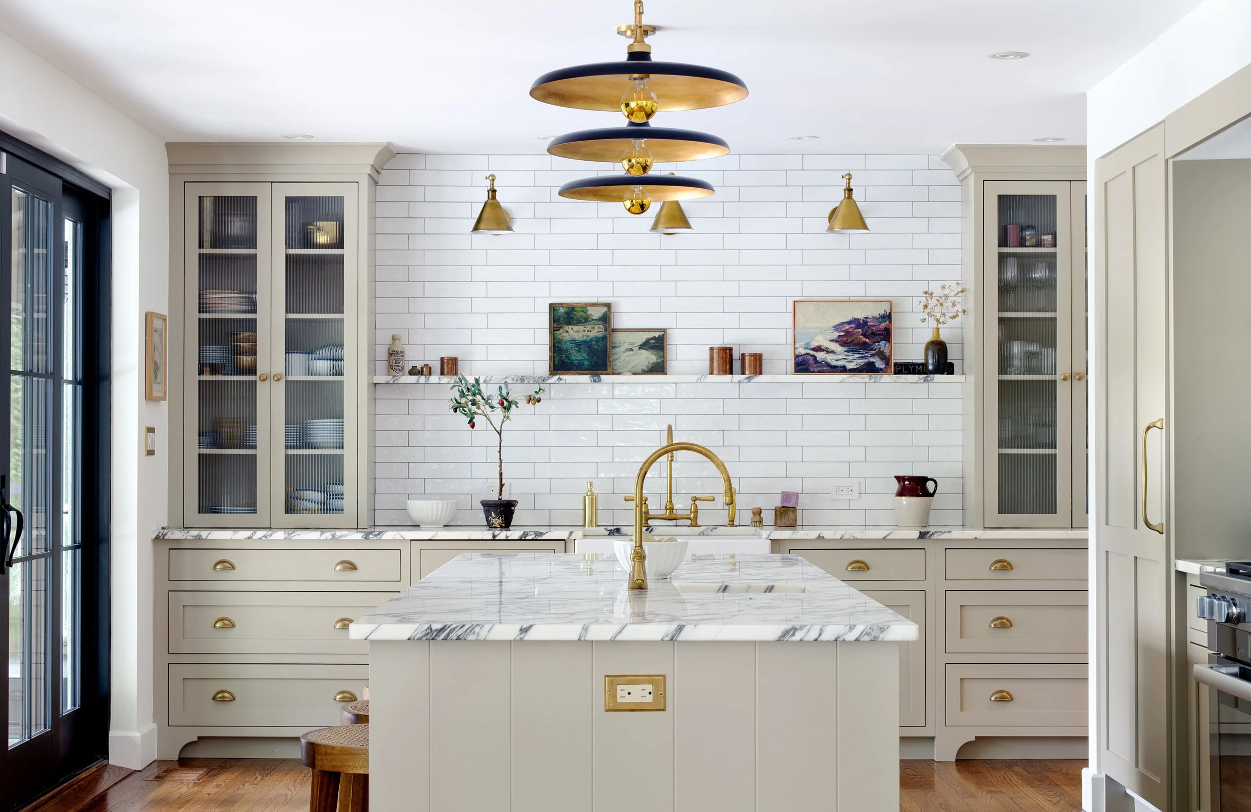 A modern kitchen with a white marble island, gold fixtures, and white cabinets with gold handles. There is a white brick backsplash and decorative paintings on a shelf. Gold and black pendant lights hang from the ceiling, and there are glass-front ca