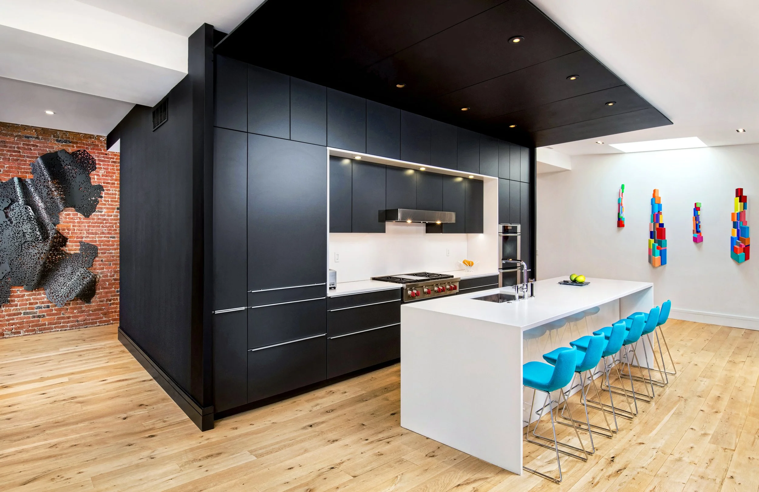 Modern kitchen with black cabinetry, white island with blue chairs, wooden flooring, brick accent wall, and colorful artwork on the white wall.