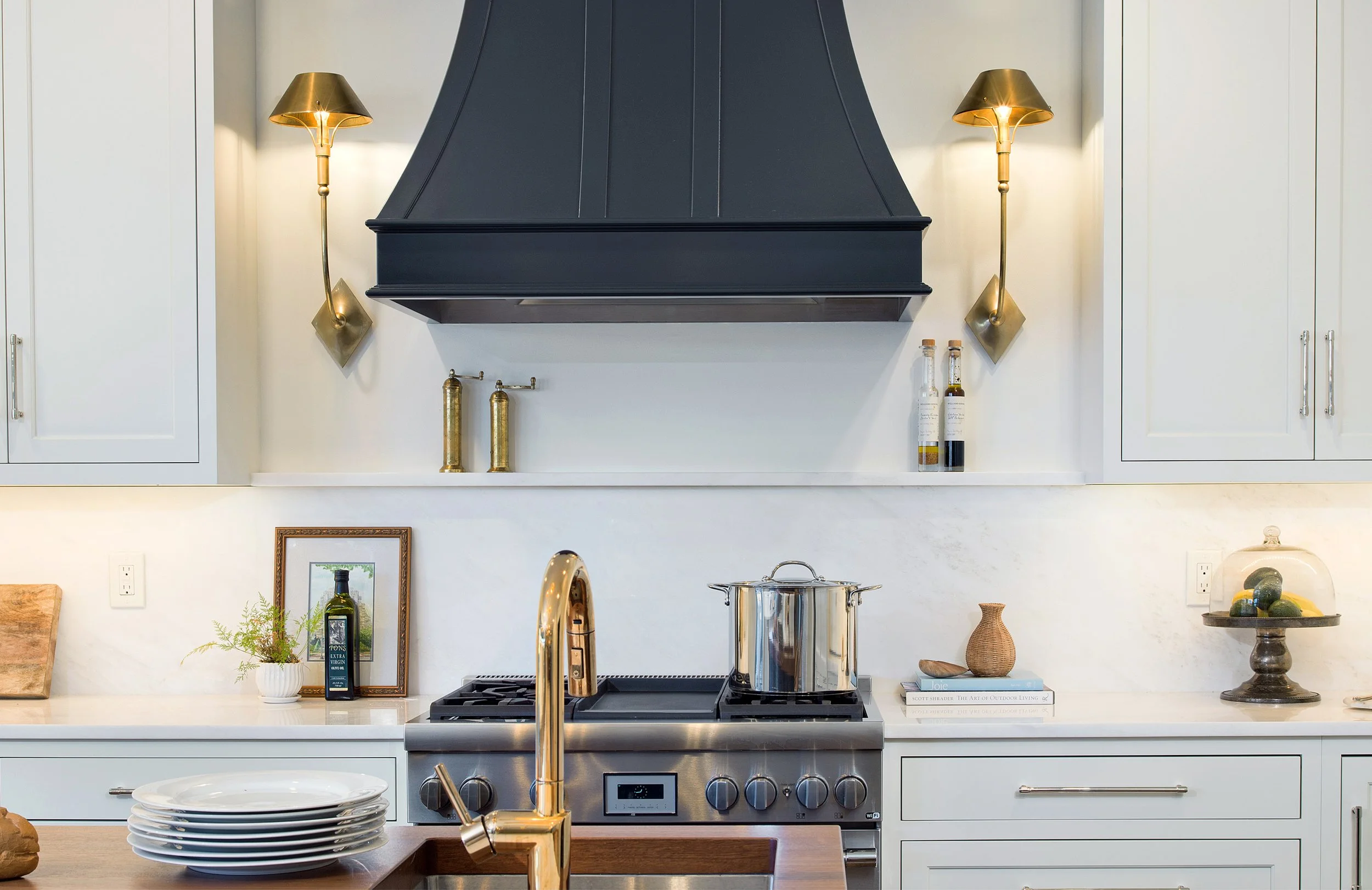 Modern kitchen with white cabinets, stainless steel stove, black range hood, gold wall sconces, and various decorative items and utensils on the countertop.