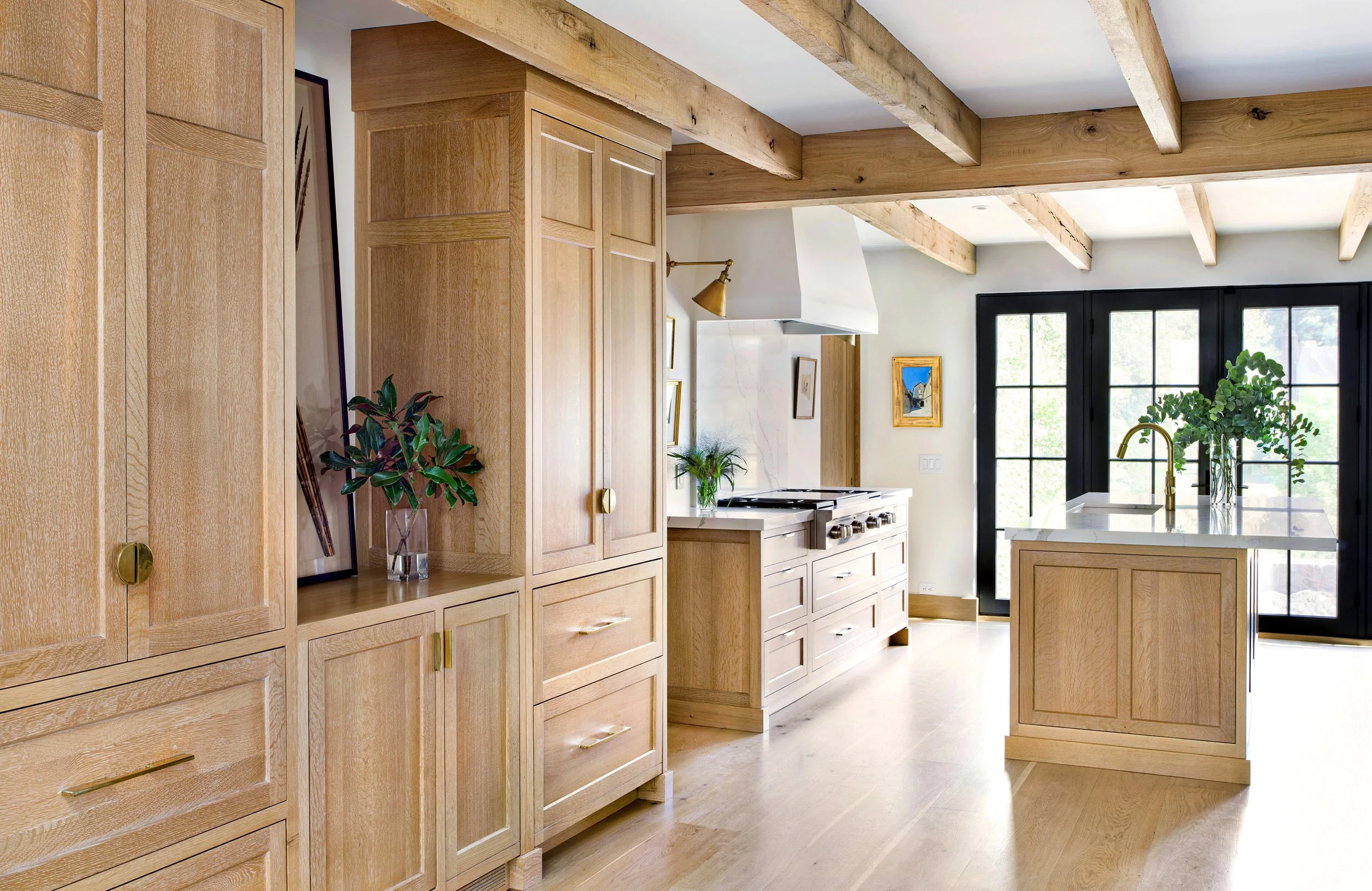Modern kitchen with wooden cabinetry, a kitchen island with plants, black-framed glass doors, and exposed wooden ceiling beams.