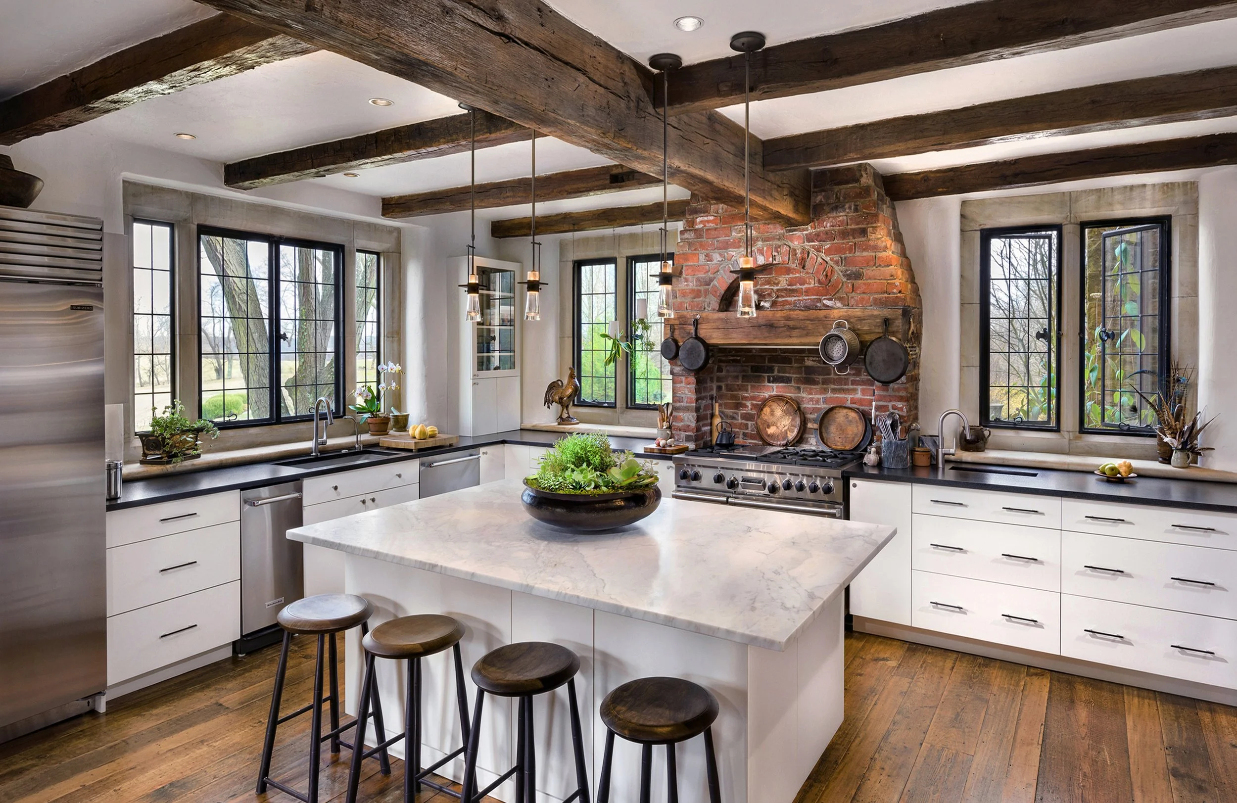 Modern Countrykitchen with white cabinets, marble island, black countertops, rustic wood beams, brick fireplace, large windows, and pendant lighting.