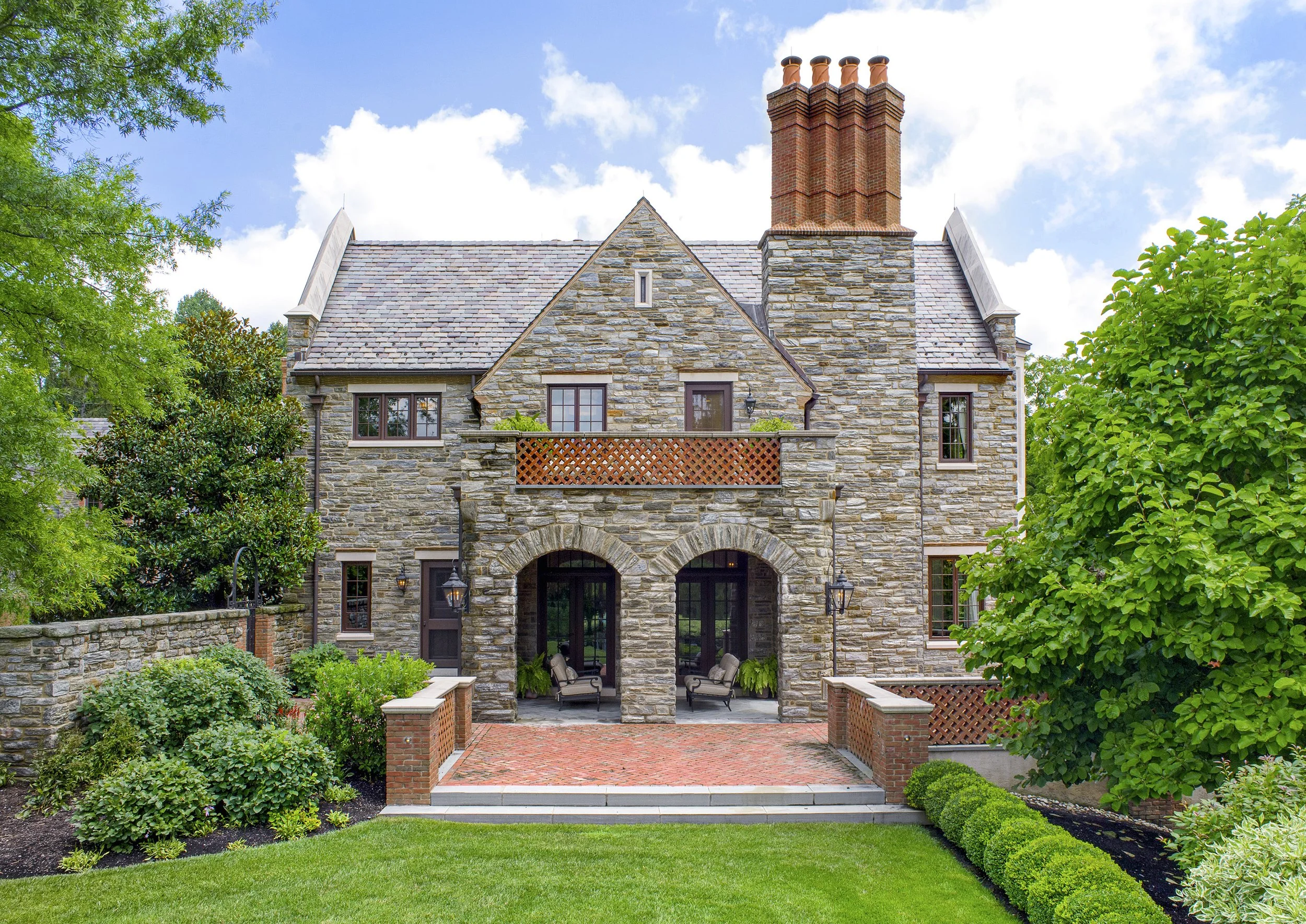 Side view of a stone house with a brick patio and chimney, lush landscape, and a blue sky with white clouds.