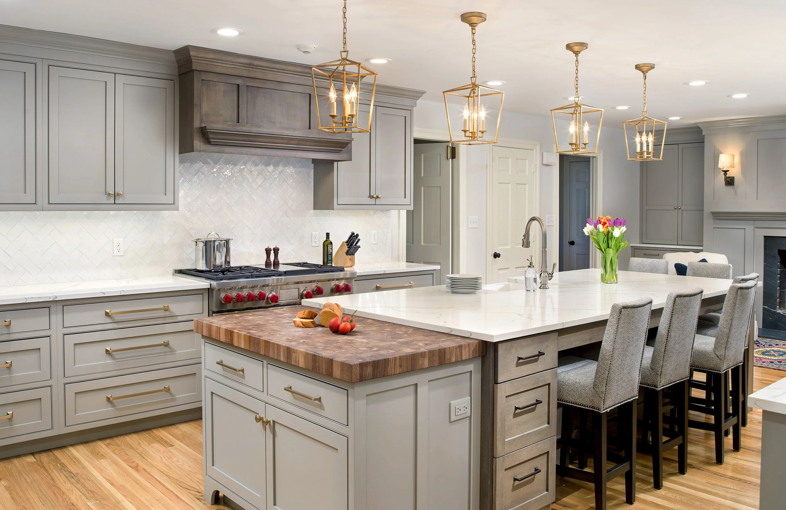 Modern kitchen with white cabinets, a large island with a white marble countertop, four upholstered bar stools, pendant lighting, a flower arrangement, and a gas stove with a wooden cutting board and fresh tomatoes.