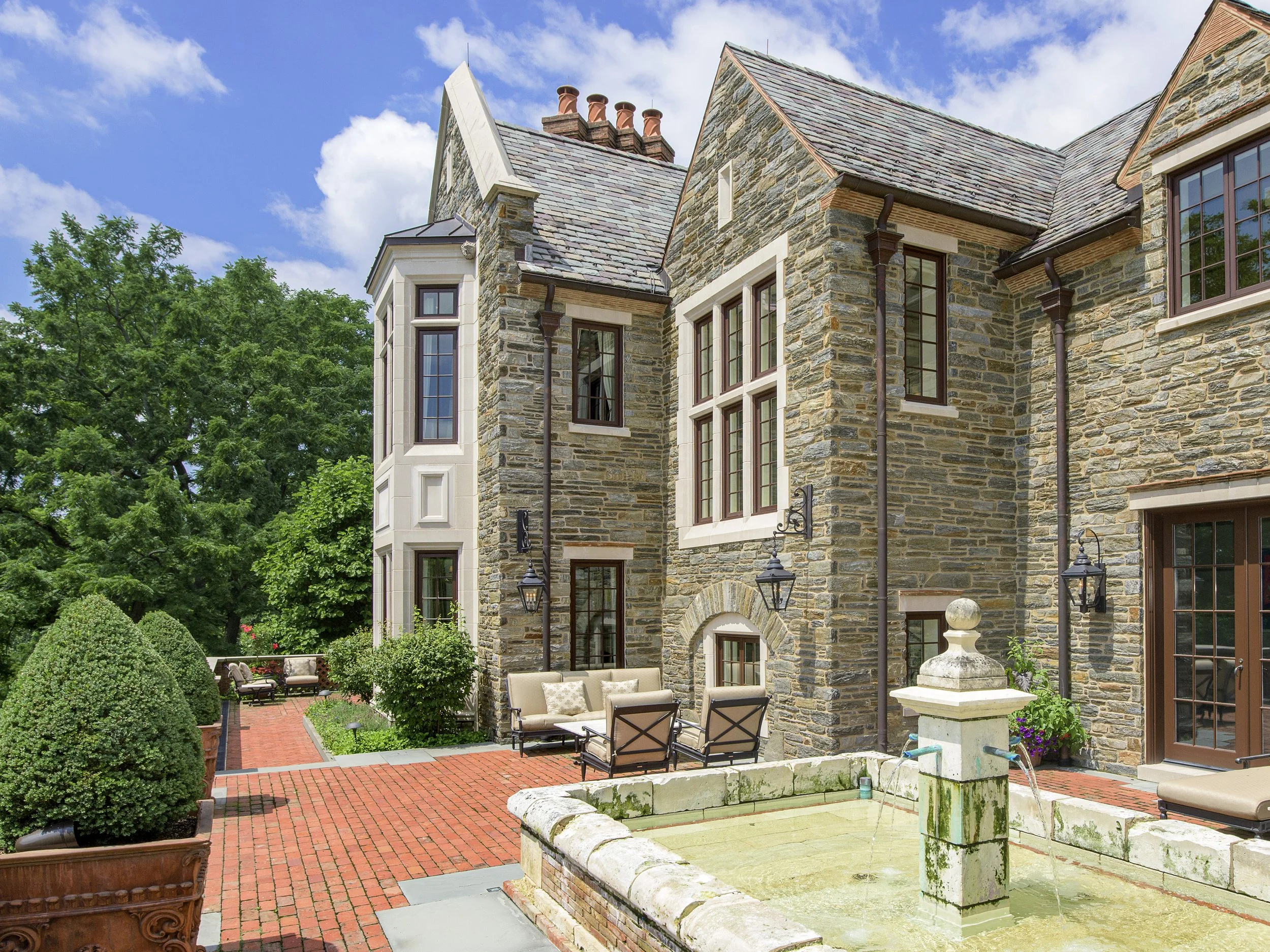 A stone house with large windows, a chimney, and a patio outdoor seating area with lawn chairs, a brick path, and a fountain in the foreground, greenery, and trees.