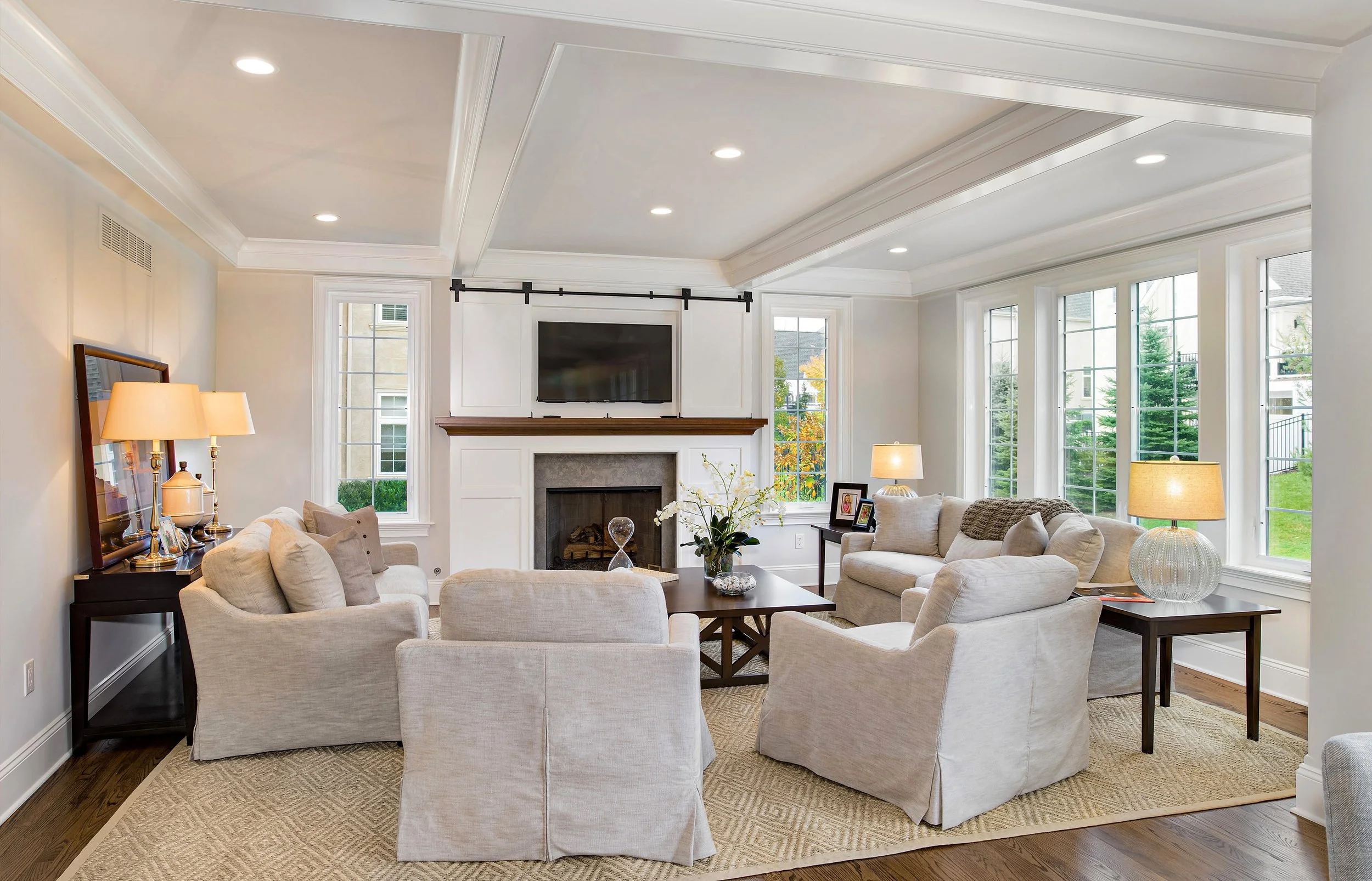 Bright living room with beige sofas, a fireplace, multiple windows, and a mounted TV above the fireplace.