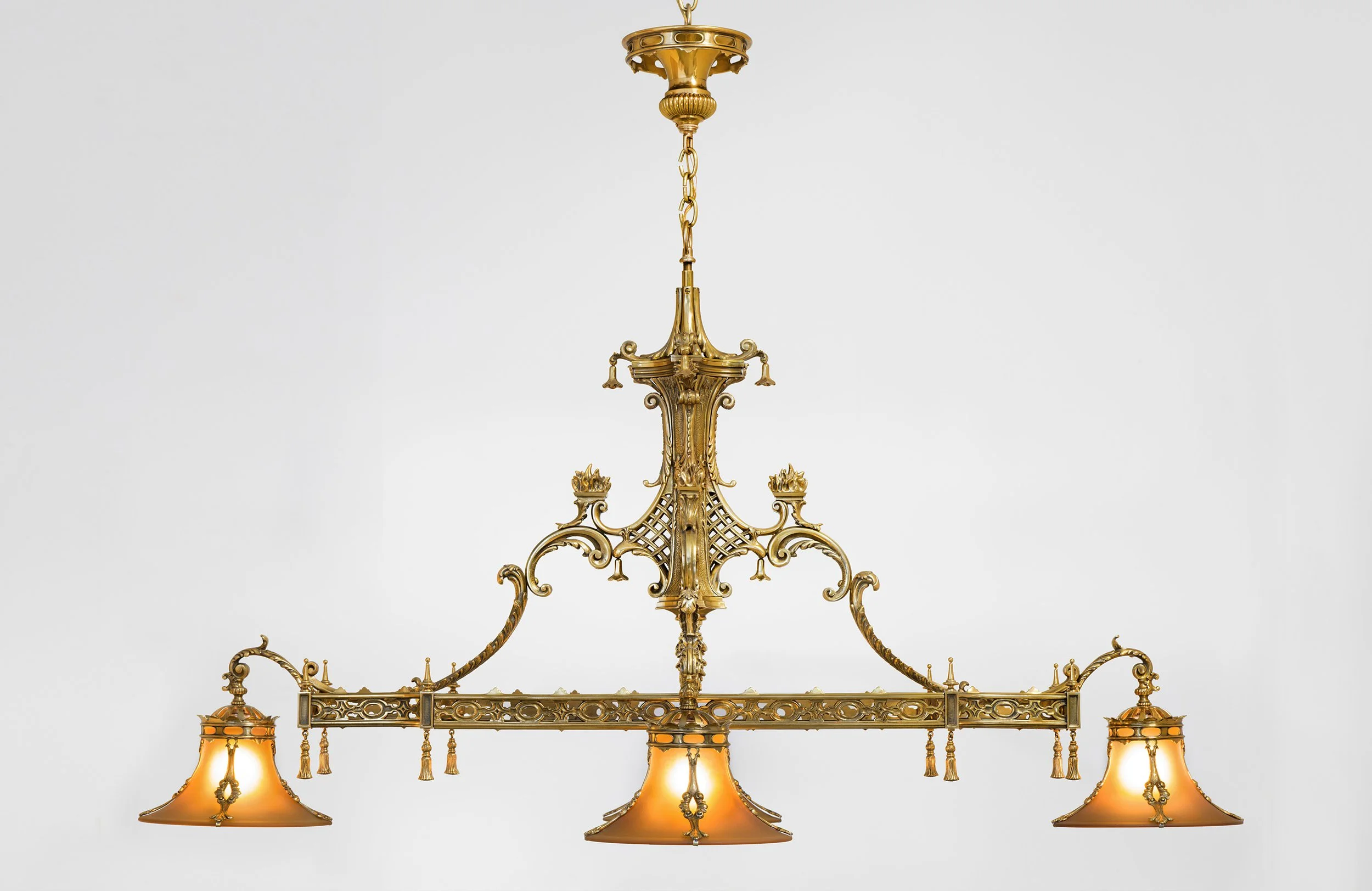 A gold chandelier with ornate decorative metalwork and amber-colored lampshades hanging from a chain and ceiling mount against a plain white background.