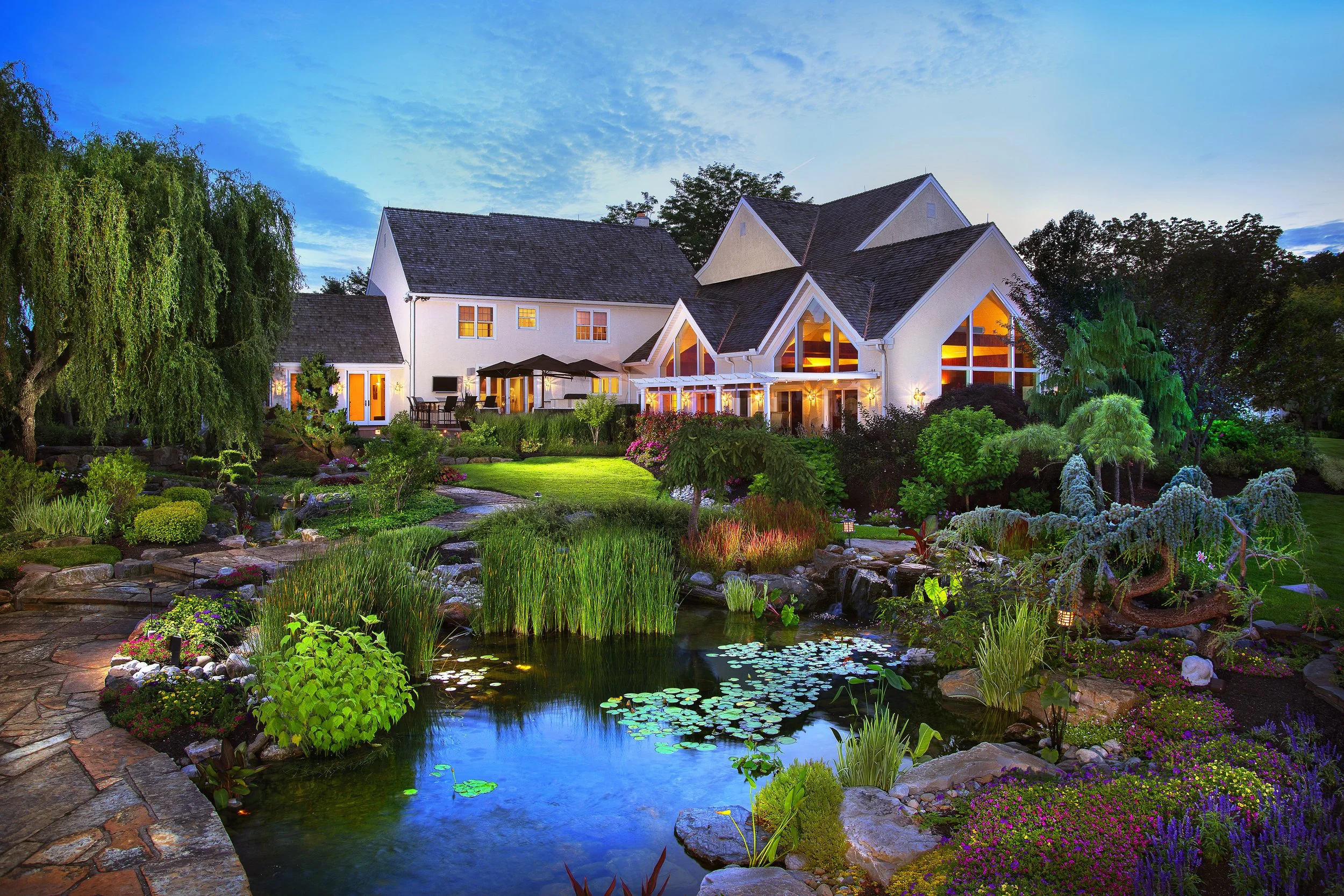 Large white house with illuminated windows, surrounded by lush gardens, stone paths, waterfalls, and a koi pond with lily pads at dusk