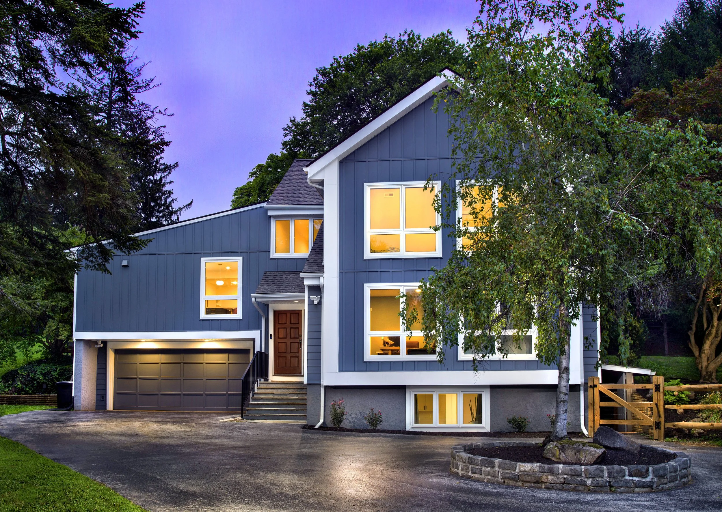 Twilight shot of a modern two-story house with dark blue exterior, large illuminated windows, a garage, and a tree in front.