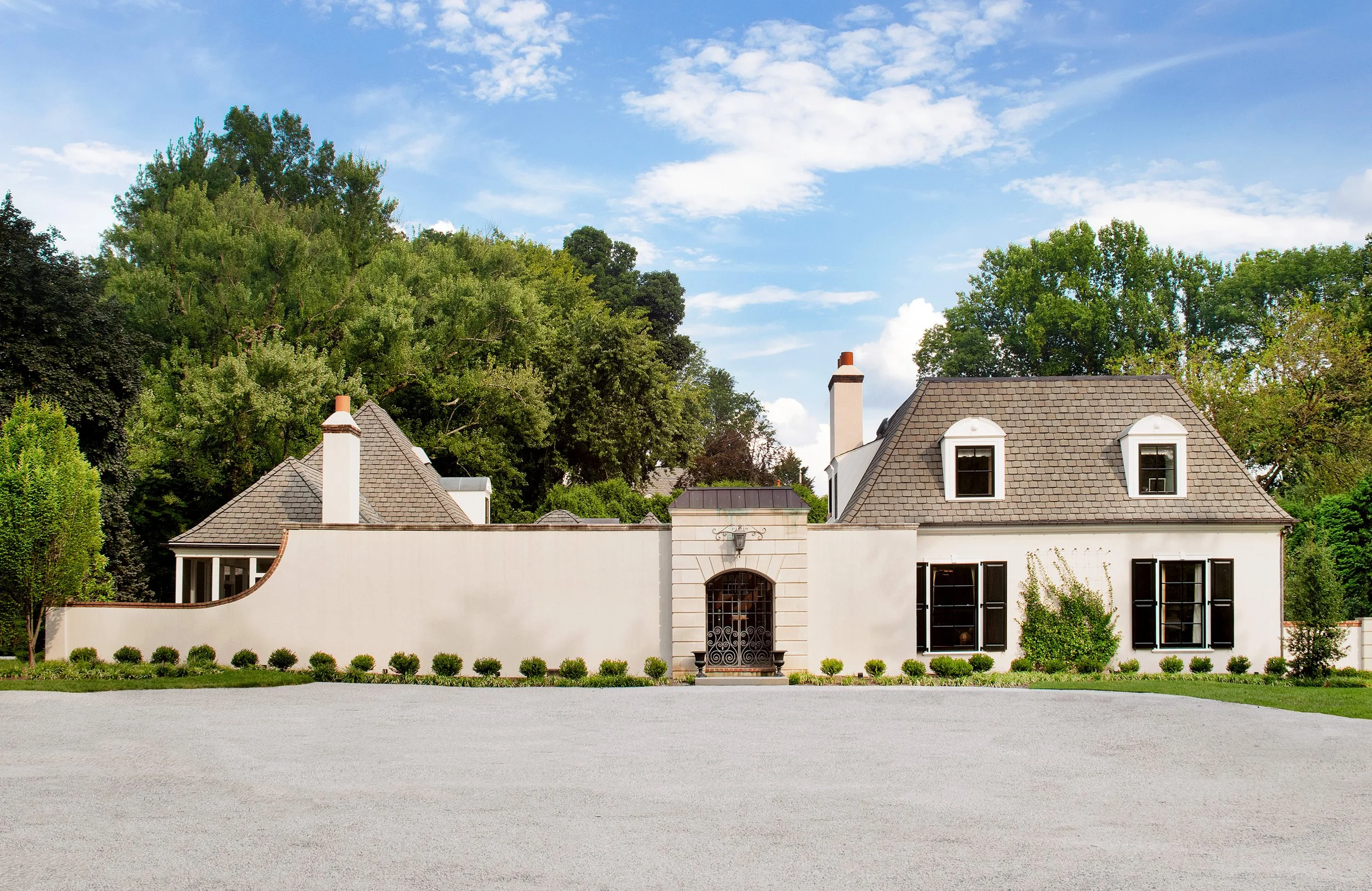 A large off-white house with black window shutters and a tiled roof. The front is surrounded with boxwood bushes that lead to a grand entry.