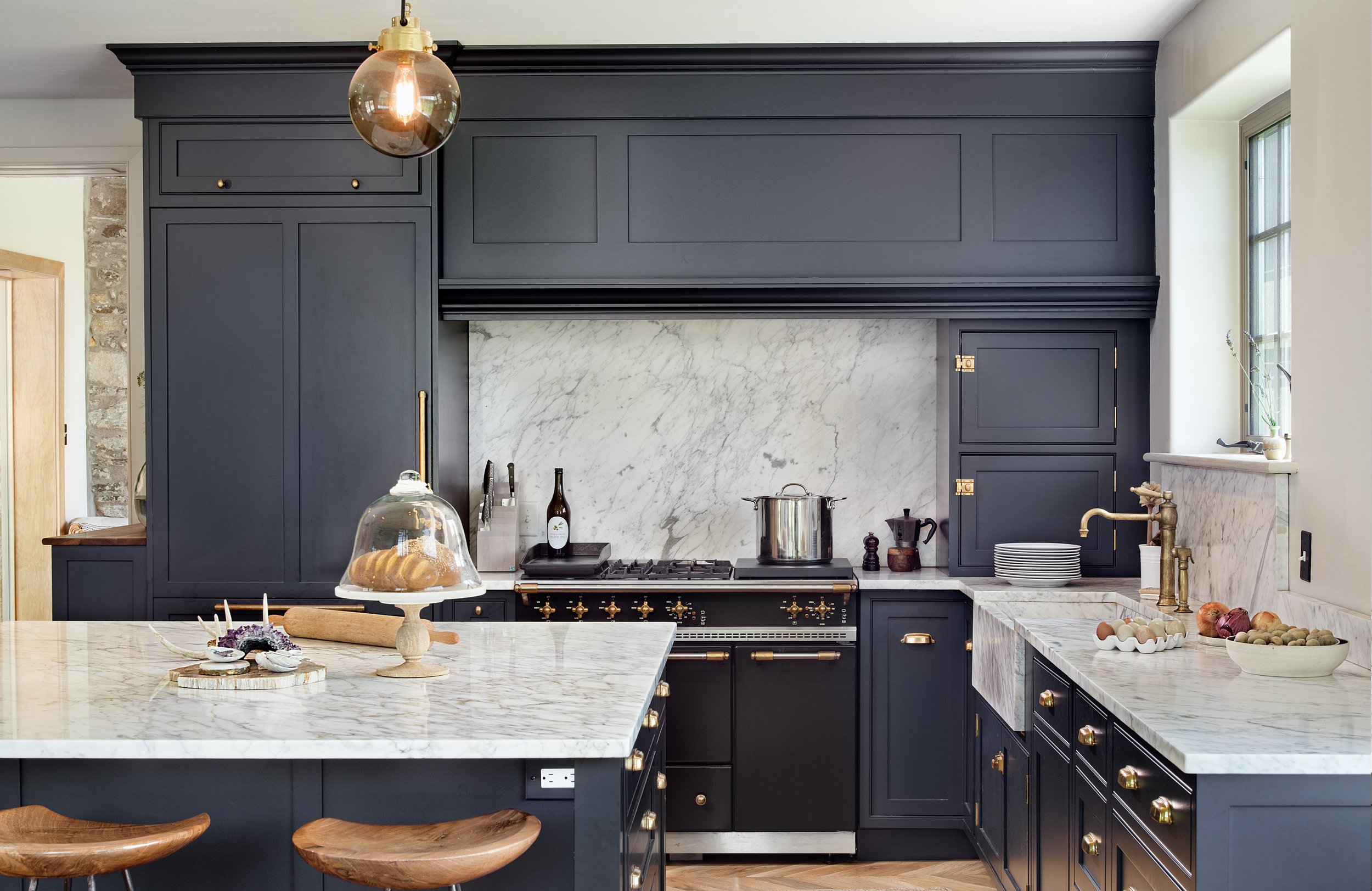 Modern kitchen with navy blue cabinets, white marble countertops, and a marble backsplash. Gold accents on hardware and fixtures, and a large island with a marble top and wooden stools. Items on countertops include a cake stand with cake under glass,