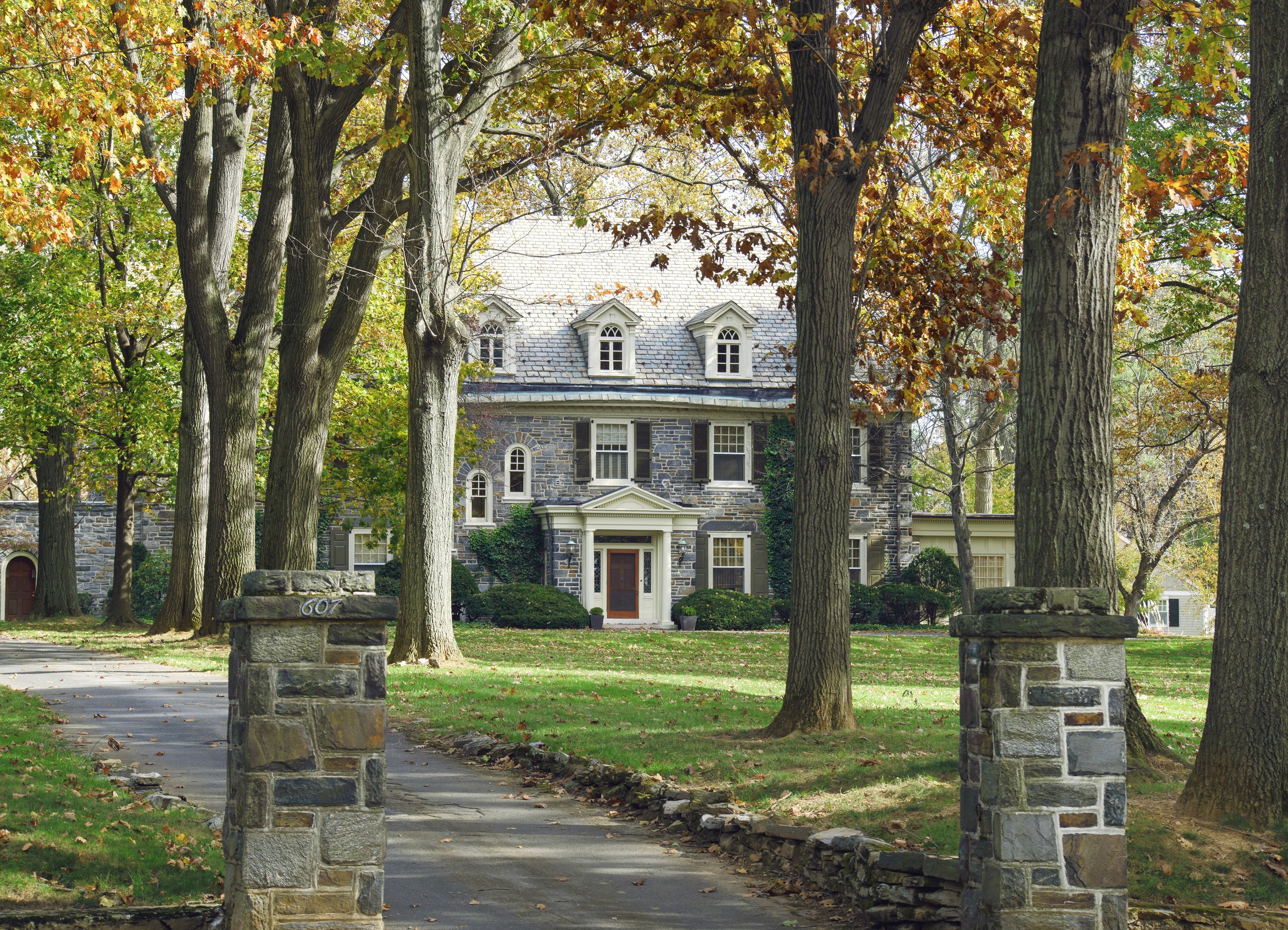 A stone house surrounded by tall trees with autumn leaves, viewed from a sidewalk with stone posts and a wooden bench.