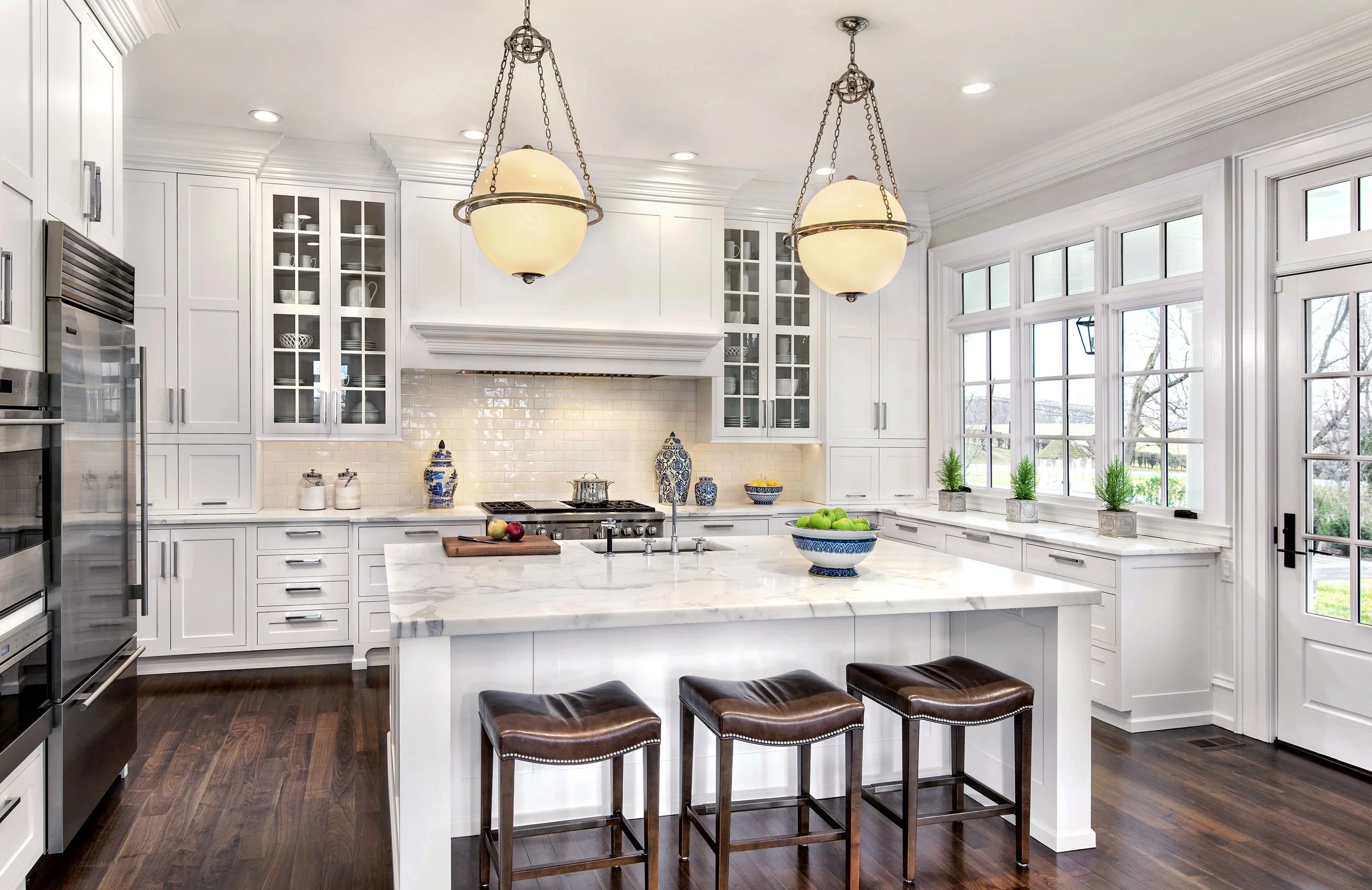 Bright kitchen with white cabinetry, marble island, dark wood flooring, large windows, and three hanging lights.