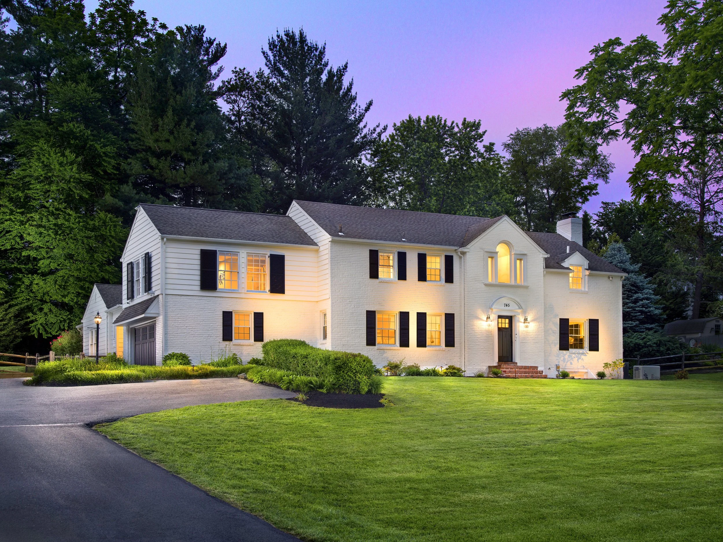 A large white two-story house with black shutters, lit from inside, with a neatly maintained green lawn and trees in the background during twilight.