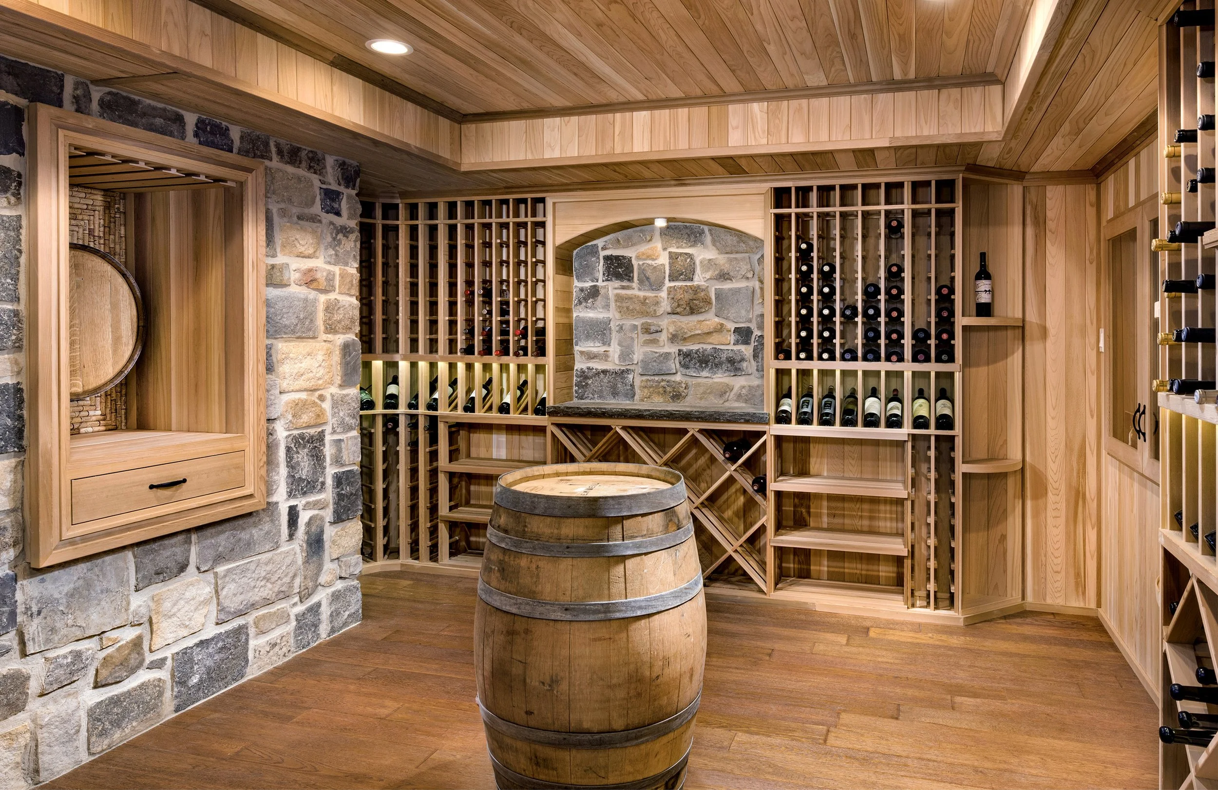 A wine cellar with wooden shelves filled with wine bottles, a stone wall accent, a wooden barrel in the center, and a wine barrel display with a pull-out drawer on the left.