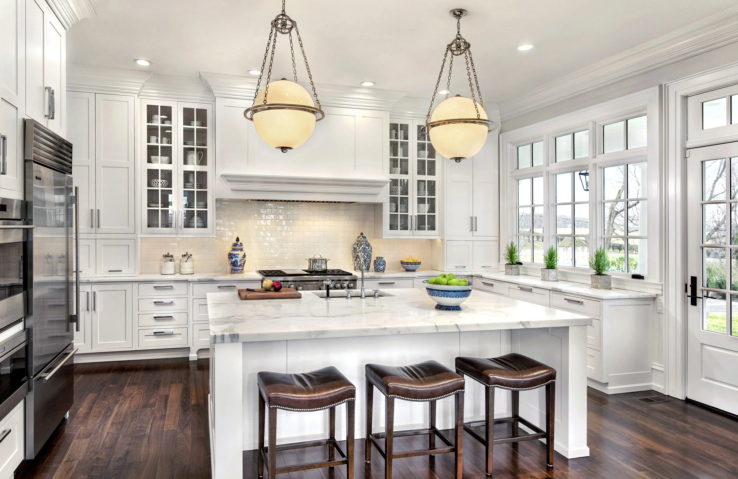 Bright kitchen with white cabinetry, marble island, dark wood flooring, large windows, and three hanging lights.