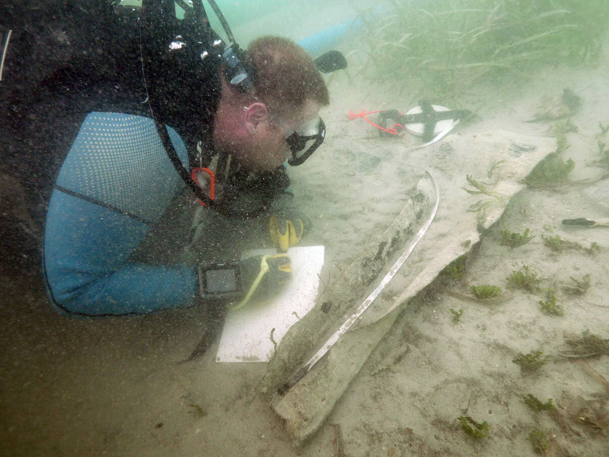 Steve Abell documenting a piece of lead sheeting which was wrapped around one of the ends of a gudgeon, possibly by the force of the explosion. Most of the square nail holes along the sides were empty of copper/bronze nails, but a few were still the